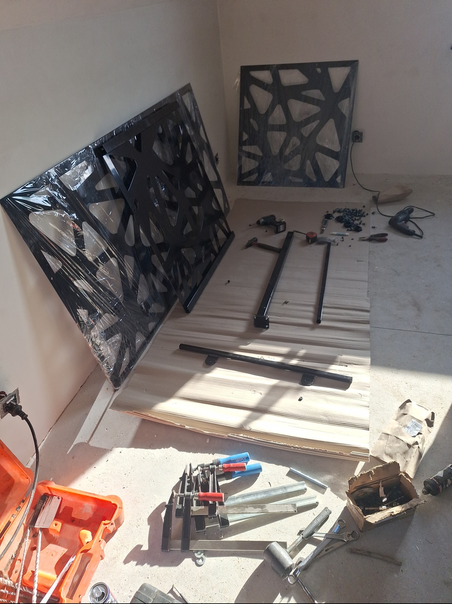 Elementy metalowe z ażurowym wzorem, częściowo w folii, leżą na drewnianej powierzchni. Widoczne narzędzia i akcesoria montażowe, sugerujące etap przygotowania do instalacji.