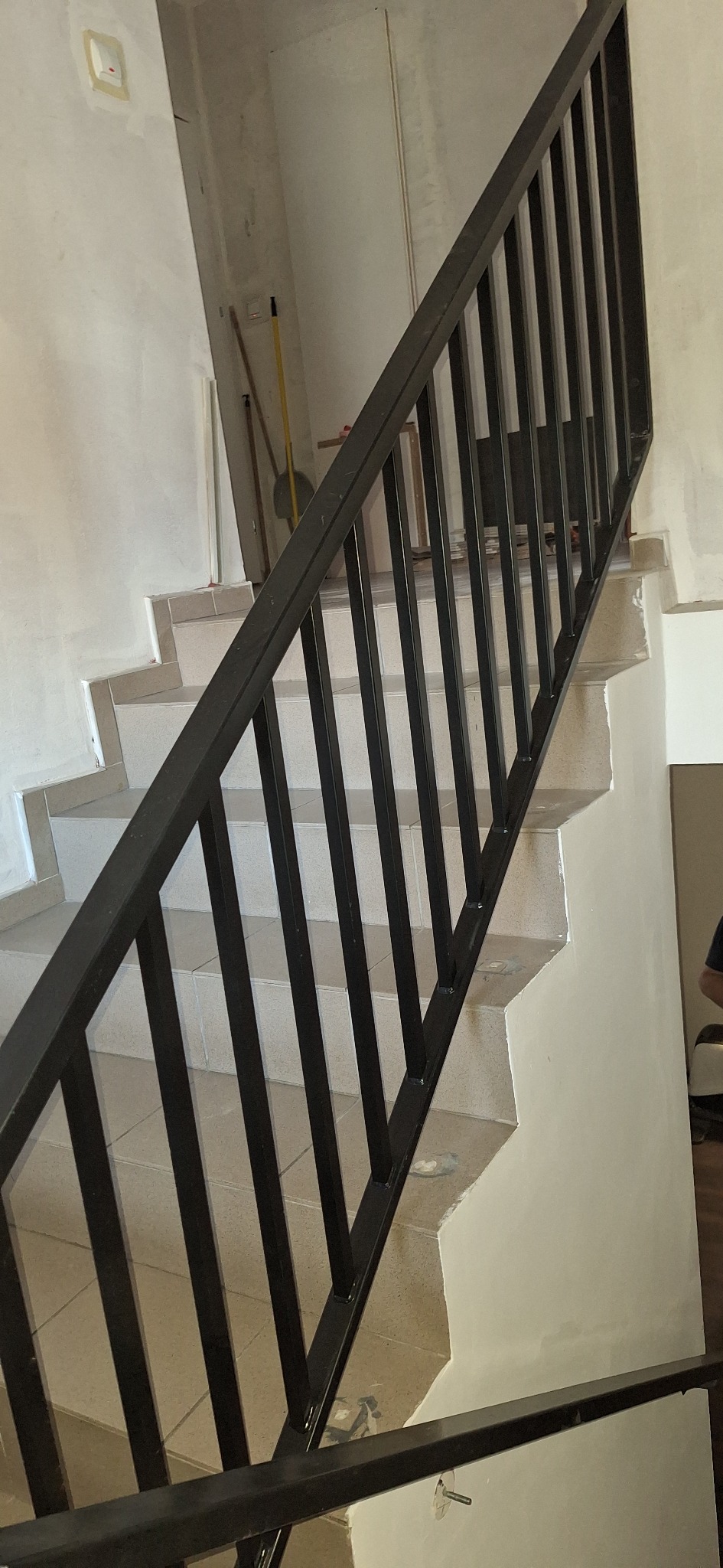 Nowoczesna, czarna poręcz schodowa z metalowymi szczeblami, zamontowana na jasnych, wykafelkowanych schodach w trakcie remontu. Widoczne niedoskonałości ścian.