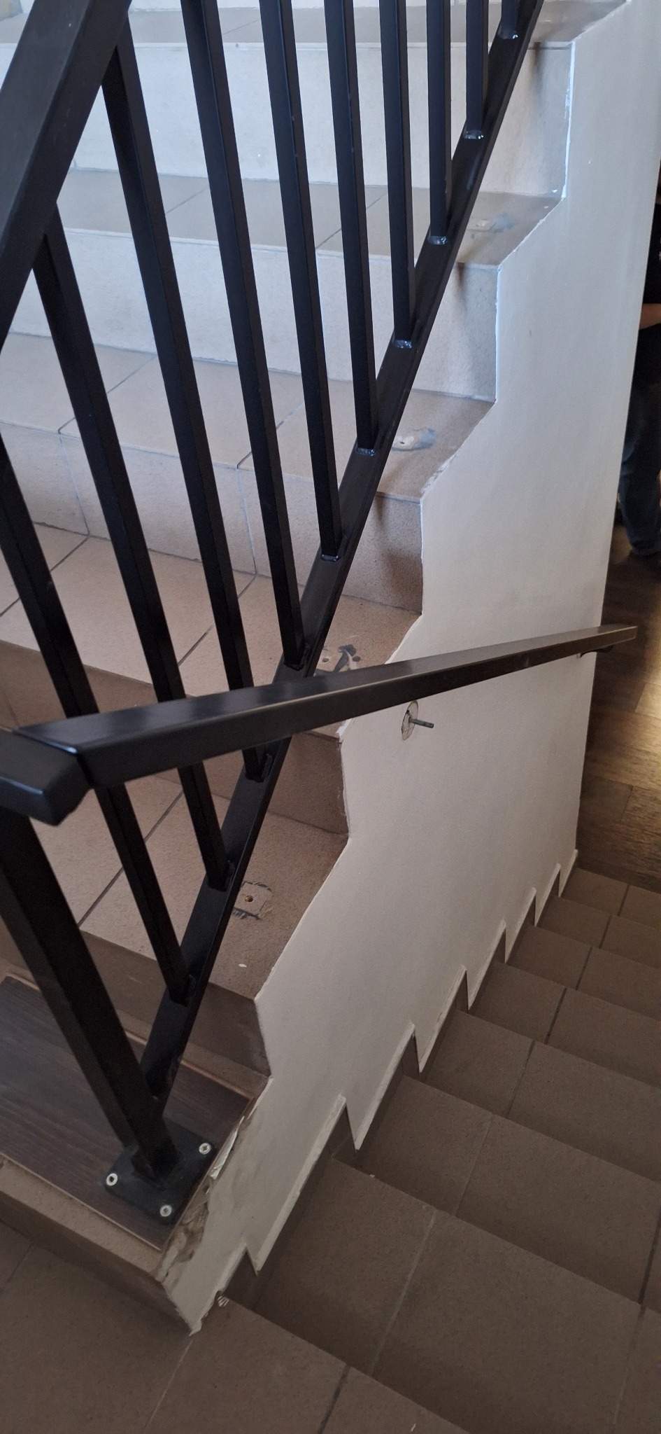 Czarne, metalowe poręcze schodowe z pionowymi szczeblami, zamocowane do ściany i stopni. Widoczne ślady montażu, płytki schodowe w odcieniach brązu.
