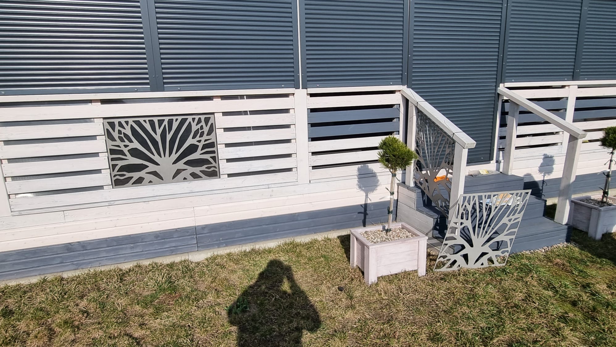 Biało-szara zabudowa balkonu z metalowymi panelami dekoracyjnymi z motywem drzewa, schody z poręczą, donice z drzewkami. Cień osoby na trawie.