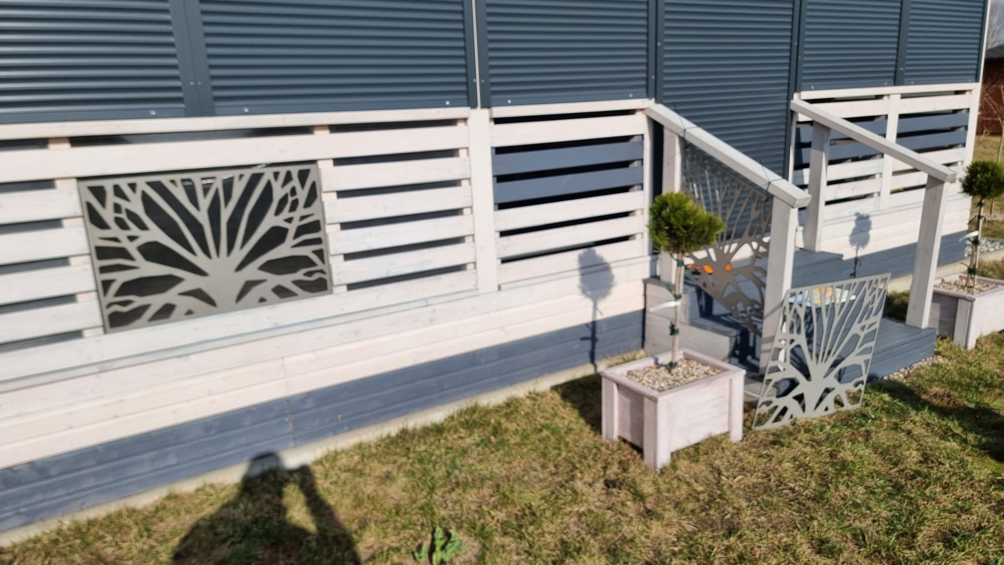 Nowoczesne ogrodzenie z drewnianymi panelami i metalowymi wstawkami z motywem drzewa, prowadzące do schodów. Dekoracyjne donice z roślinami.