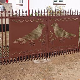 Iskrownia Grzegorz Michnowski - Brązowa brama wjazdowa z ażurowym wzorem dwóch gołębi, otoczona metalowym ogrodzeniem z dekoracyjnymi grotami. Widoczne świeże prace ziemne.