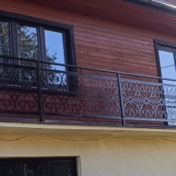 Iskrownia Grzegorz Michnowski - Czarna, metalowa balustrada balkonowa z dekoracyjnym, ażurowym wzorem na tle drewnianej elewacji domu. Widoczne okna z ciemnymi ramami i numer domu '90'.