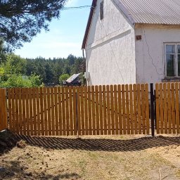 Iskrownia Grzegorz Michnowski - Nowe, drewniane ogrodzenie sztachetowe z bramką, świeżo zamontowane na posesji z domem. Widoczne fundamenty i częściowo wykopana ziemia.