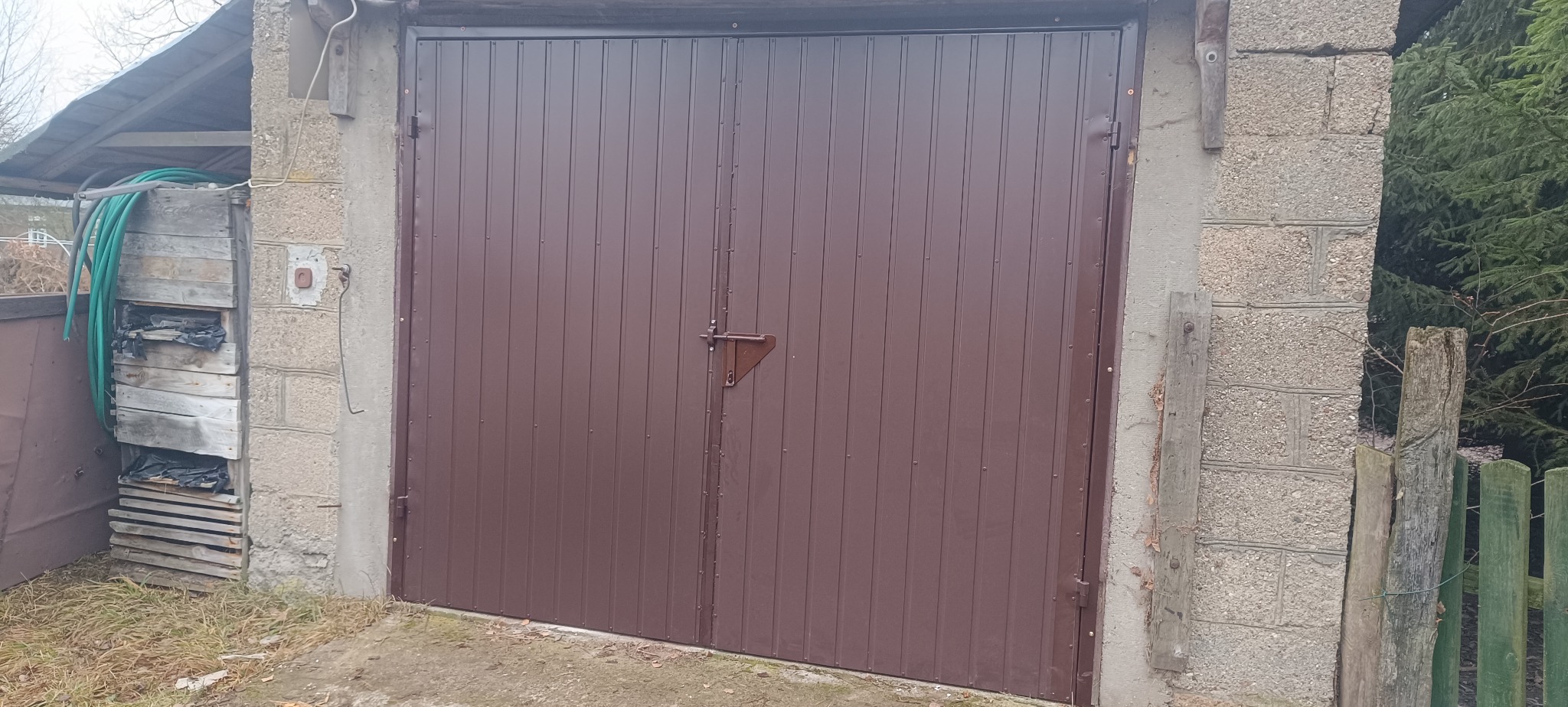 Brązowa, dwuskrzydłowa brama garażowa z prostym zamkiem, osadzona w betonowych słupkach. Obok widoczna drewniana konstrukcja i fragment zielonego ogrodzenia.