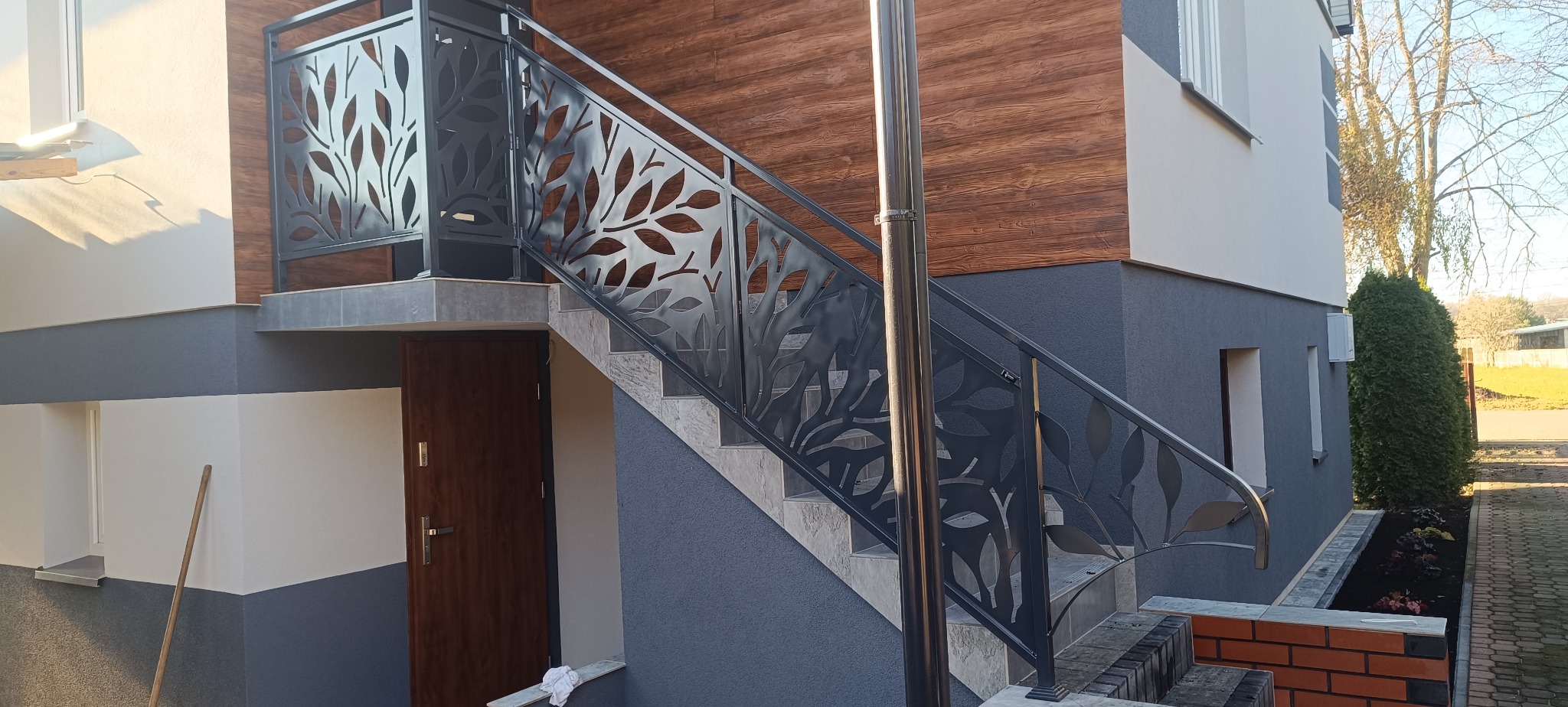 Nowoczesna balustrada schodowa z motywem liści, wykonana z metalu w kolorze grafitowym. Schody prowadzą do drzwi wejściowych budynku z drewnianą elewacją.