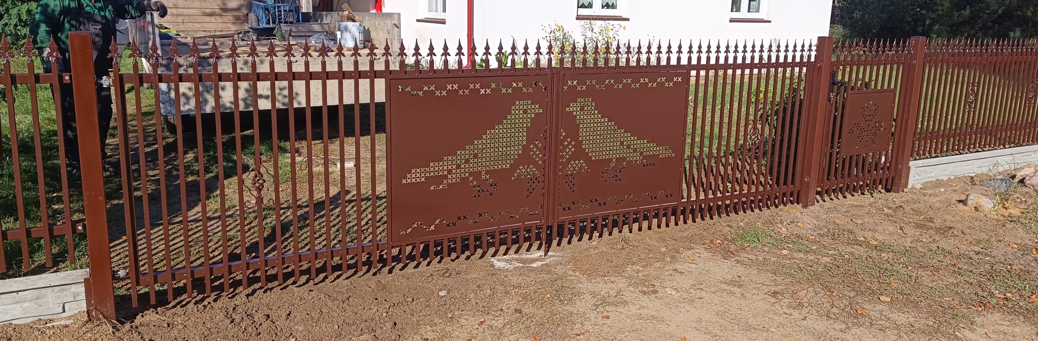 Brązowa brama wjazdowa z ażurowym wzorem dwóch gołębi, otoczona metalowym ogrodzeniem z dekoracyjnymi grotami. Widoczne świeże prace ziemne.