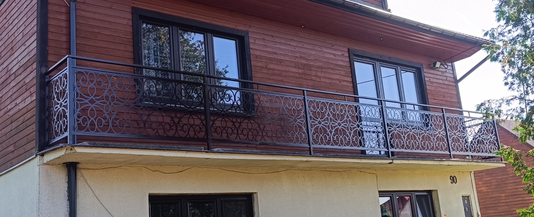 Czarna, metalowa balustrada balkonowa z dekoracyjnym, ażurowym wzorem na tle drewnianej elewacji domu. Widoczne okna z ciemnymi ramami i numer domu '90'.