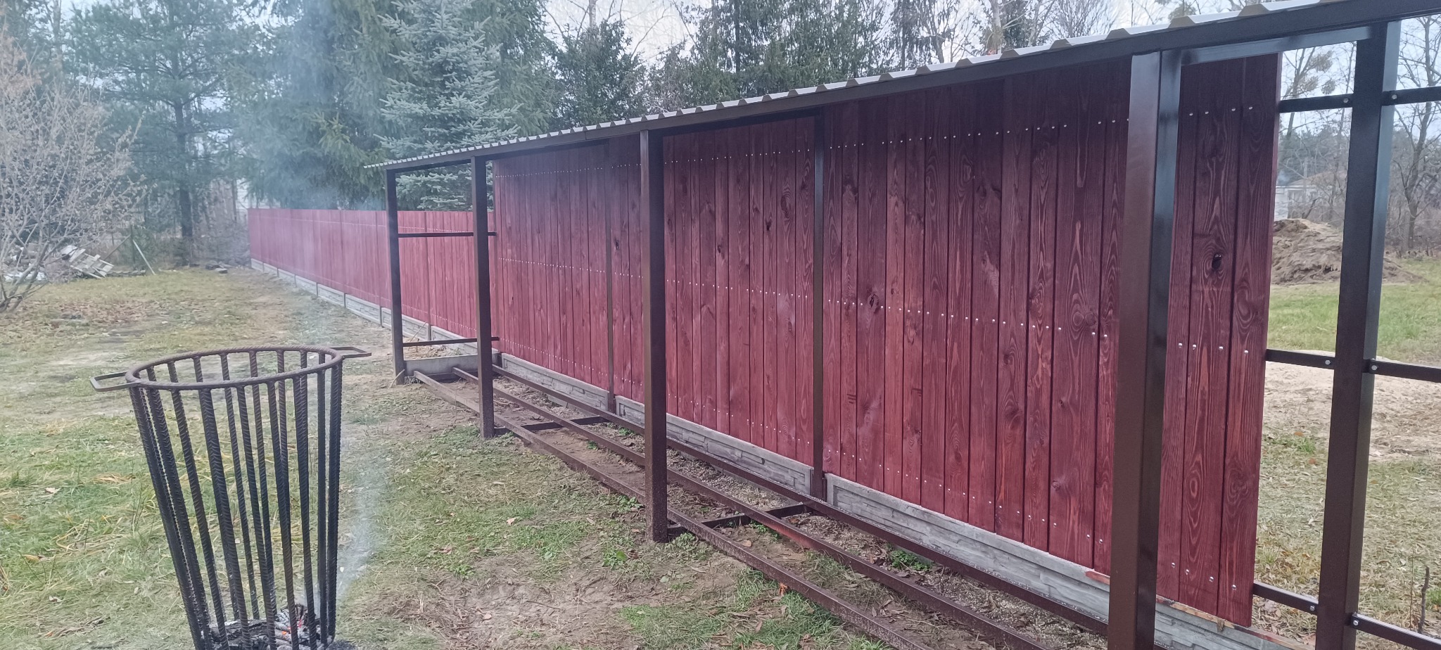Częściowo zadaszone, bordowe ogrodzenie z drewnianych desek i metalowych słupków, w trakcie montażu, z widoczną konstrukcją podstawy. W tle drzewa i palenisko.