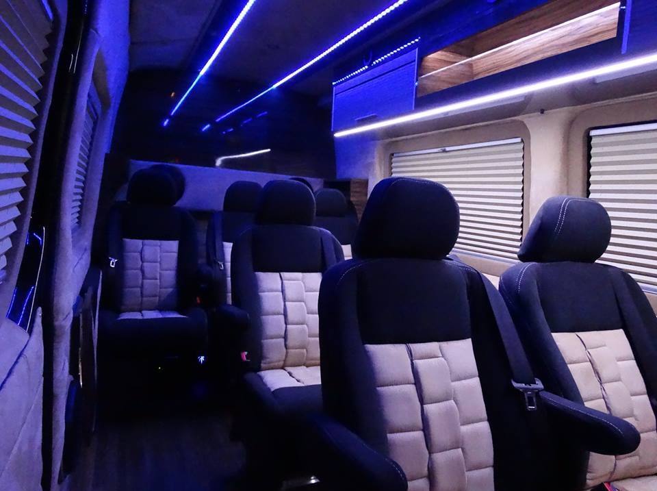 Wnętrze luksusowego busa z czarno-beżowymi fotelami, oświetlonego niebieskimi neonami. Widok z tyłu pojazdu na fotele pasażerów i okna z żaluzjami.