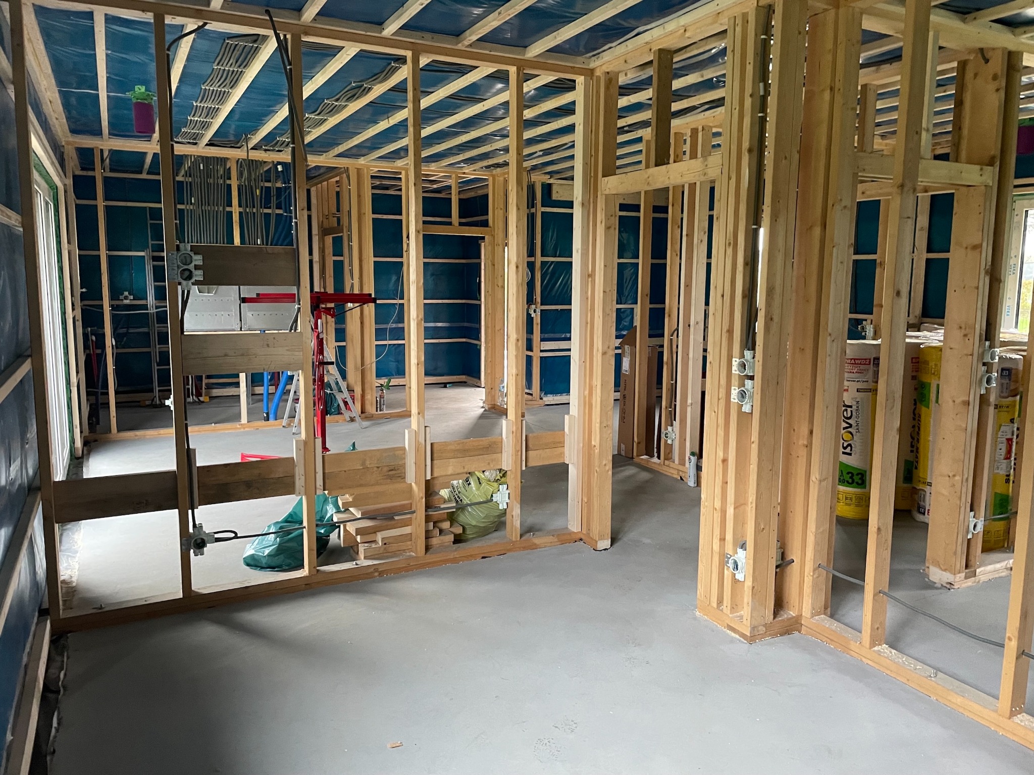 Wnętrze w trakcie budowy z drewnianą konstrukcją szkieletową, widoczne przewody elektryczne i puszki instalacyjne na ścianach. Niebieska folia na suficie.