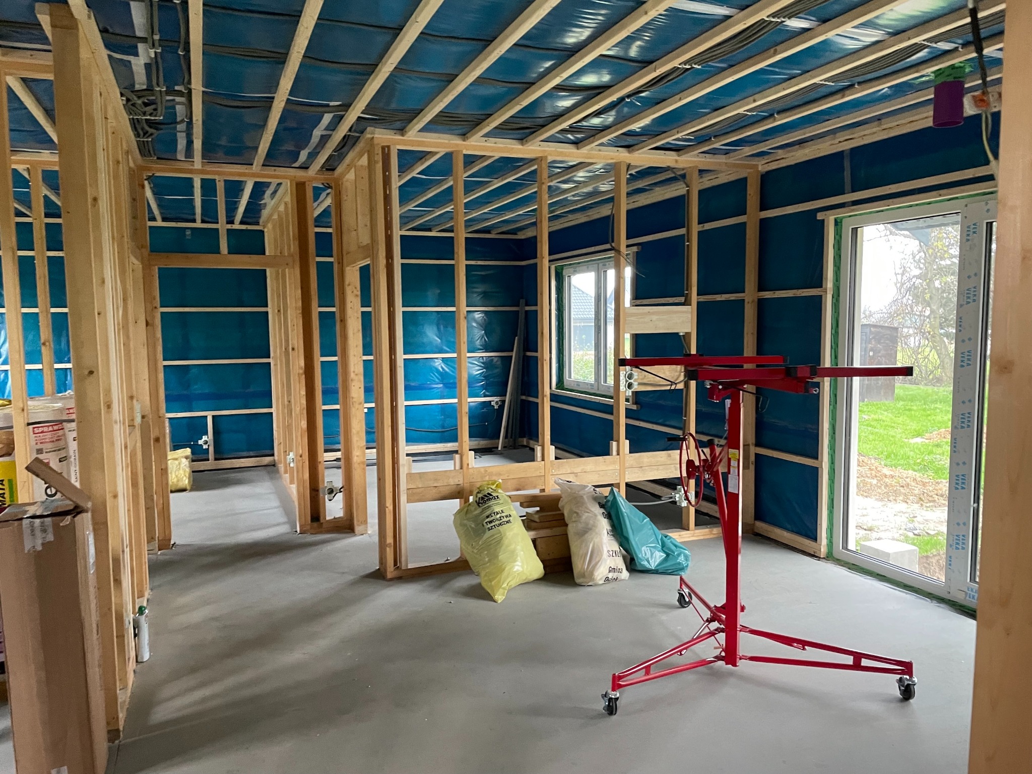 Wnętrze w trakcie prac wykończeniowych: drewniana konstrukcja ścian, instalacje pod sufitem, podnośnik do płyt gipsowych i worki z materiałami budowlanymi.