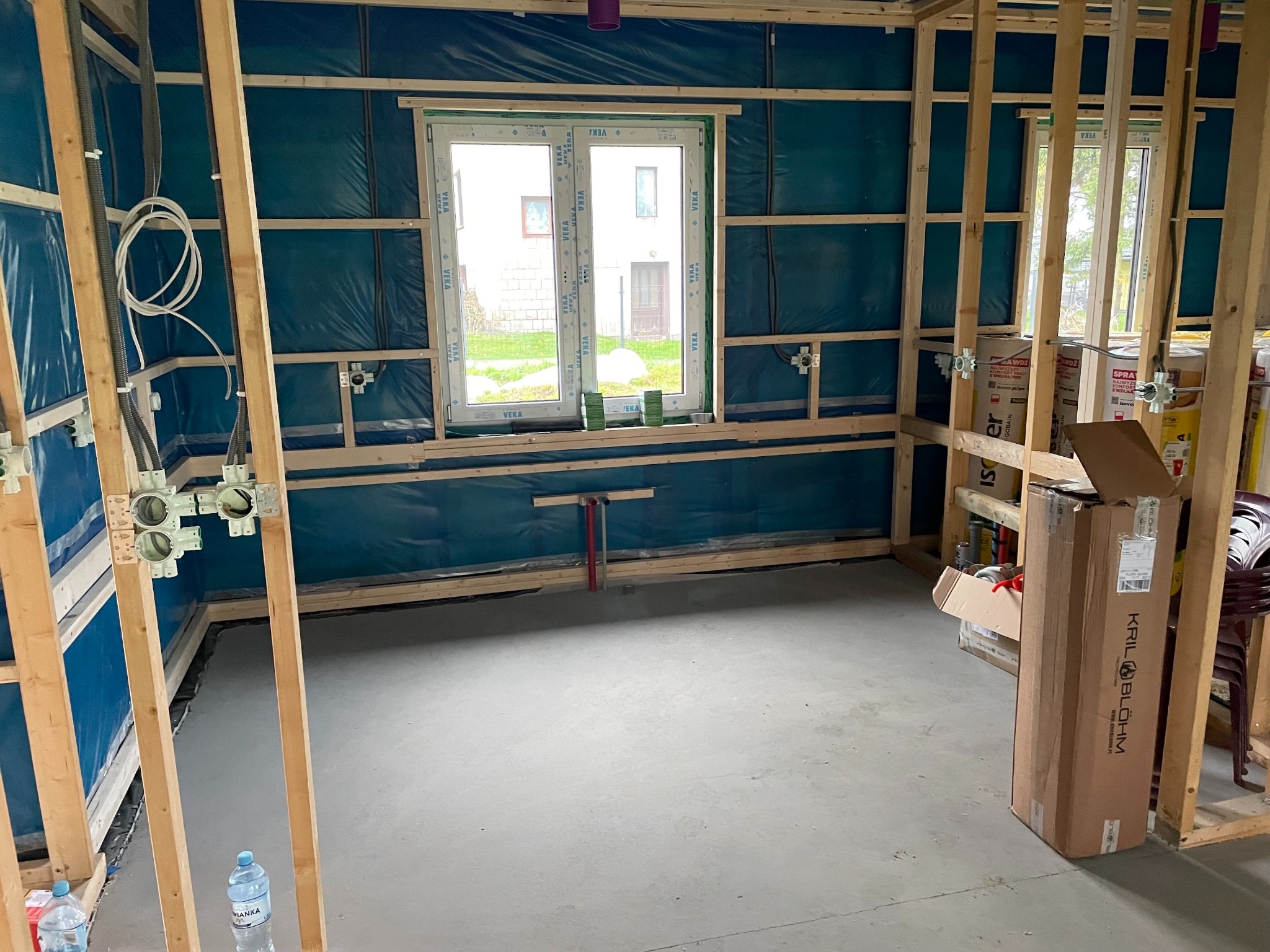 Surowy stan wnętrza z drewnianą konstrukcją ścian, instalacją elektryczną w puszkach i przewodach. Widoczne okno i podłoże betonowe. Etap prac wykończeniowych.