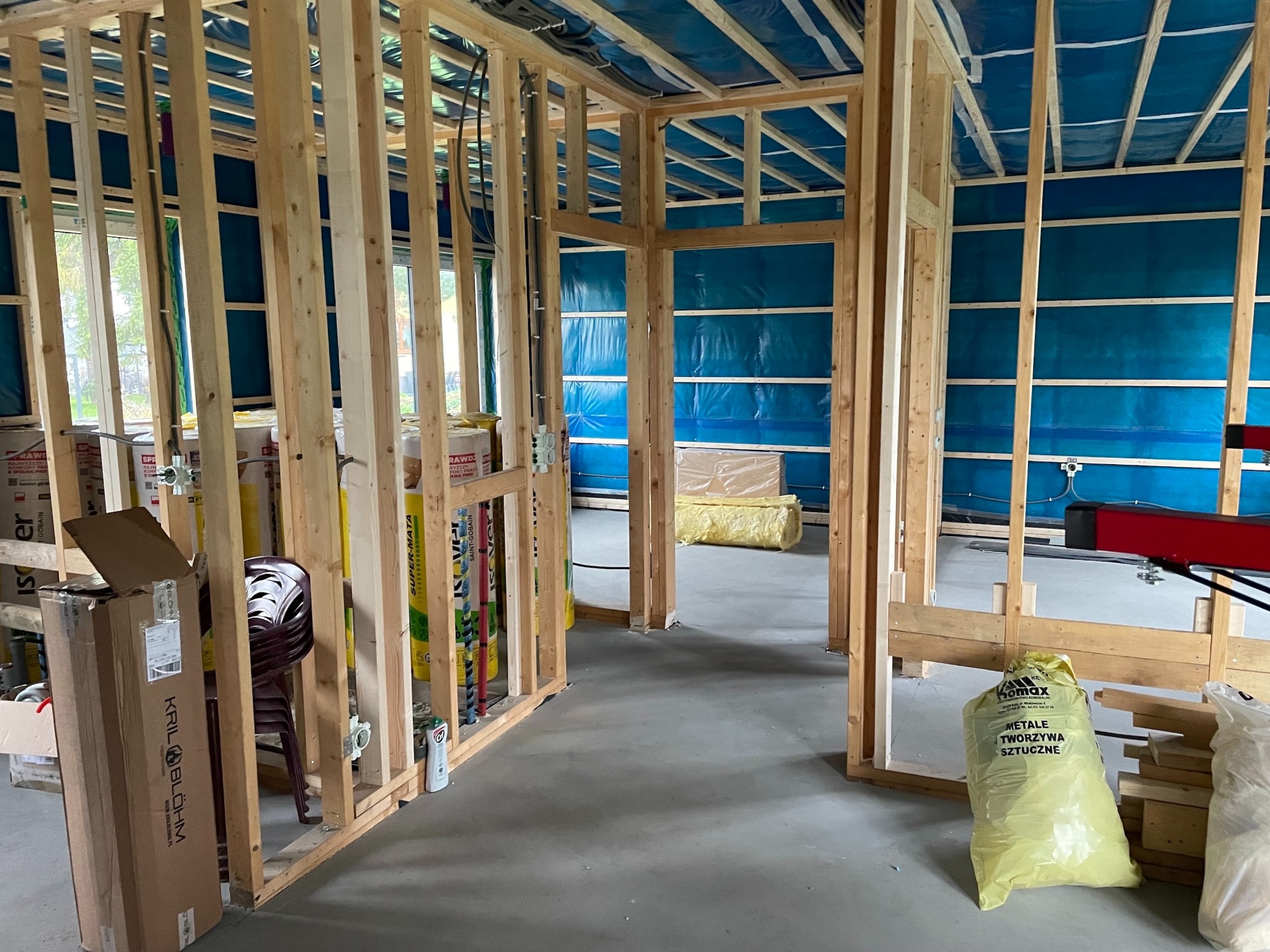 Wnętrze w trakcie budowy: drewniana konstrukcja ścian, niebieska folia izolacyjna, widoczne przewody elektryczne. Etap prac przed wykończeniem.