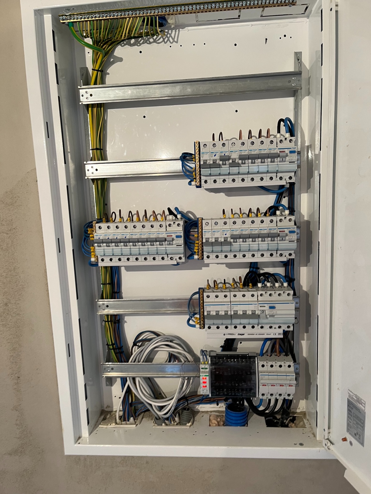 Rozdzielnica elektryczna w białej obudowie, widoczne zabezpieczenia, przewody i listwy montażowe. Instalacja w trakcie realizacji, widoczne kable w różnych kolorach.