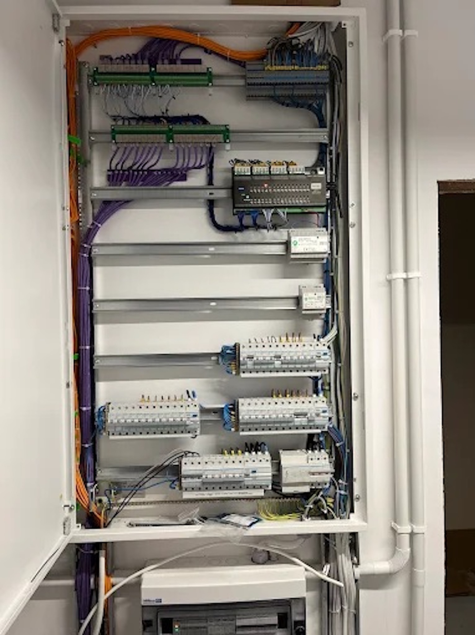 Otwarta skrzynka elektryczna z licznymi przewodami w kolorach pomarańczowym i fioletowym, zamontowana na ścianie wewnątrz budynku. Widoczne zabezpieczenia i listwy.