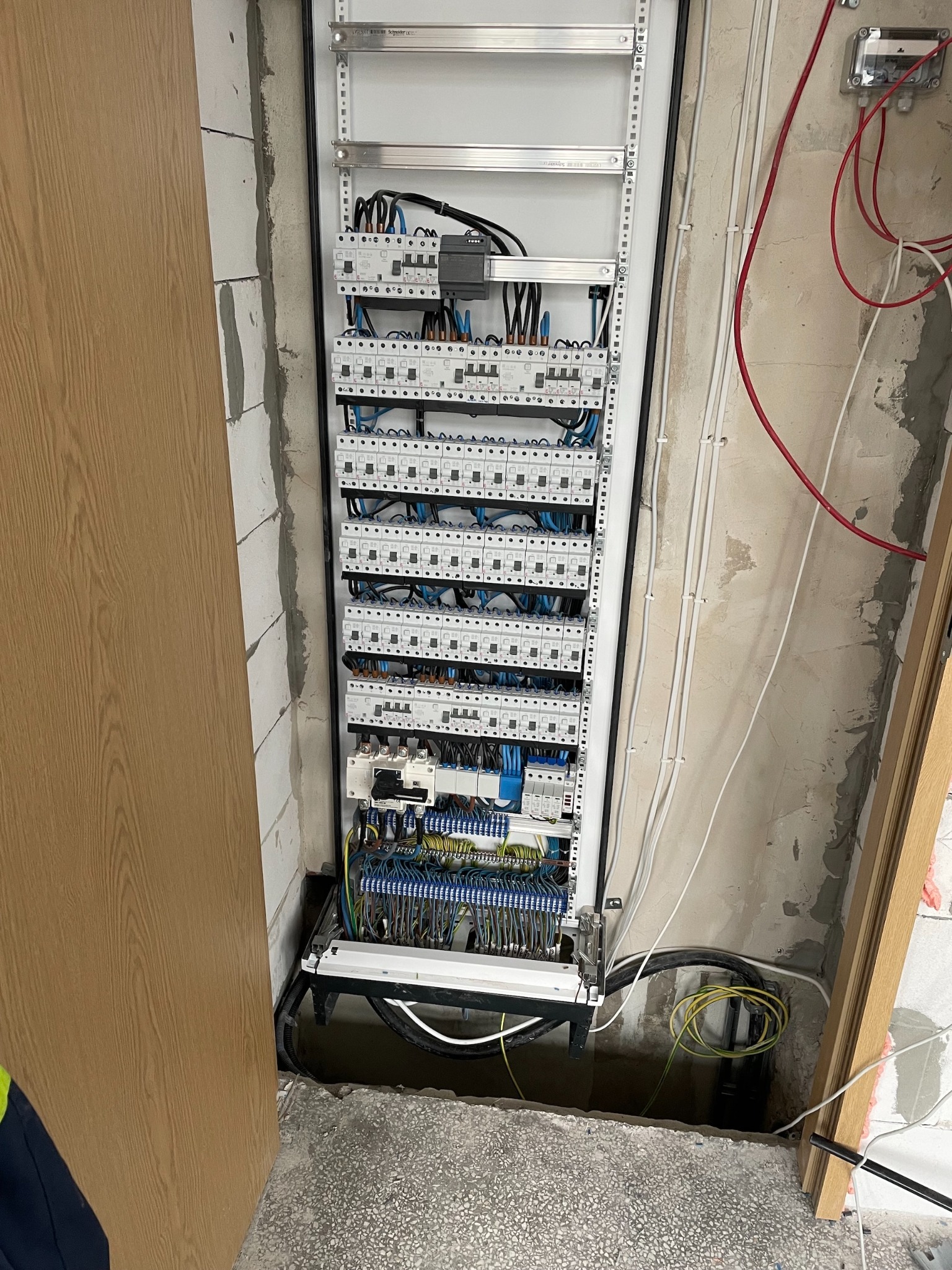 Rozdzielnica elektryczna z licznymi zabezpieczeniami i przewodami, zamontowana w surowym wnętrzu. Widoczne kable i elementy instalacji na ścianie.