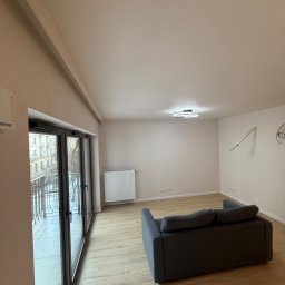 Globi-Bud - Jasne wnętrze po remoncie: parkiet, szara sofa, okno balkonowe z widokiem na miasto. Na ścianie widoczne niedokończone instalacje elektryczne.