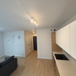 Globi-Bud - Jasne, nowocześnie wykończone wnętrze mieszkania z drewnianą podłogą, białymi ścianami i zabudową kuchenną. Widoczne elementy dekoracyjne i oświetlenie.