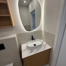 Globi-Bud - Nowoczesna łazienka po remoncie: owalne lustro z podświetleniem LED, biała umywalka nablatowa na drewnianej szafce z marmurowym blatem, szare płytki.