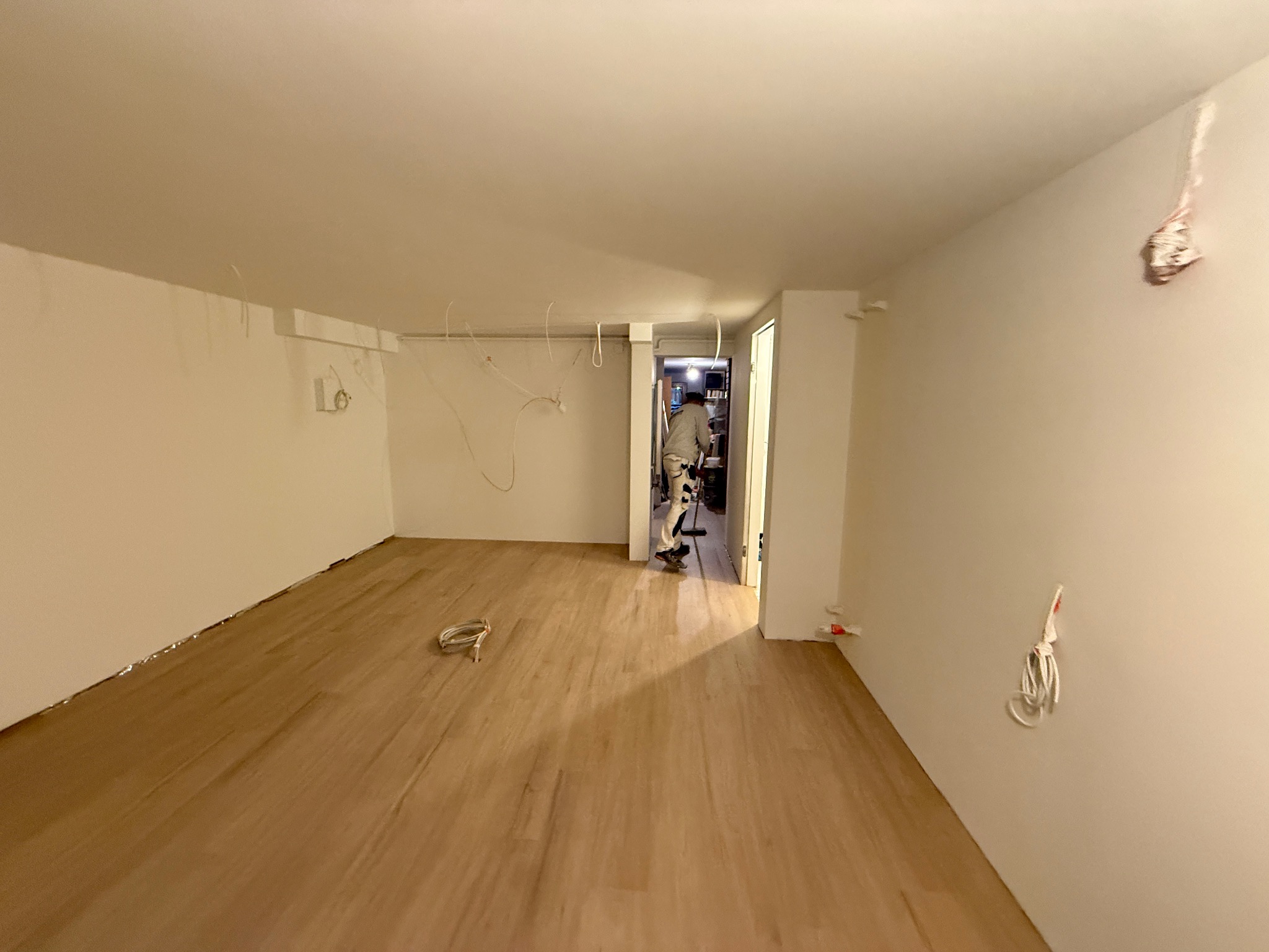 Jasne, puste pomieszczenie z drewnianą podłogą, przygotowane do malowania. Widoczne kable wystające ze ścian i sufitu. Pracownik w oddali przygotowuje się do pracy.