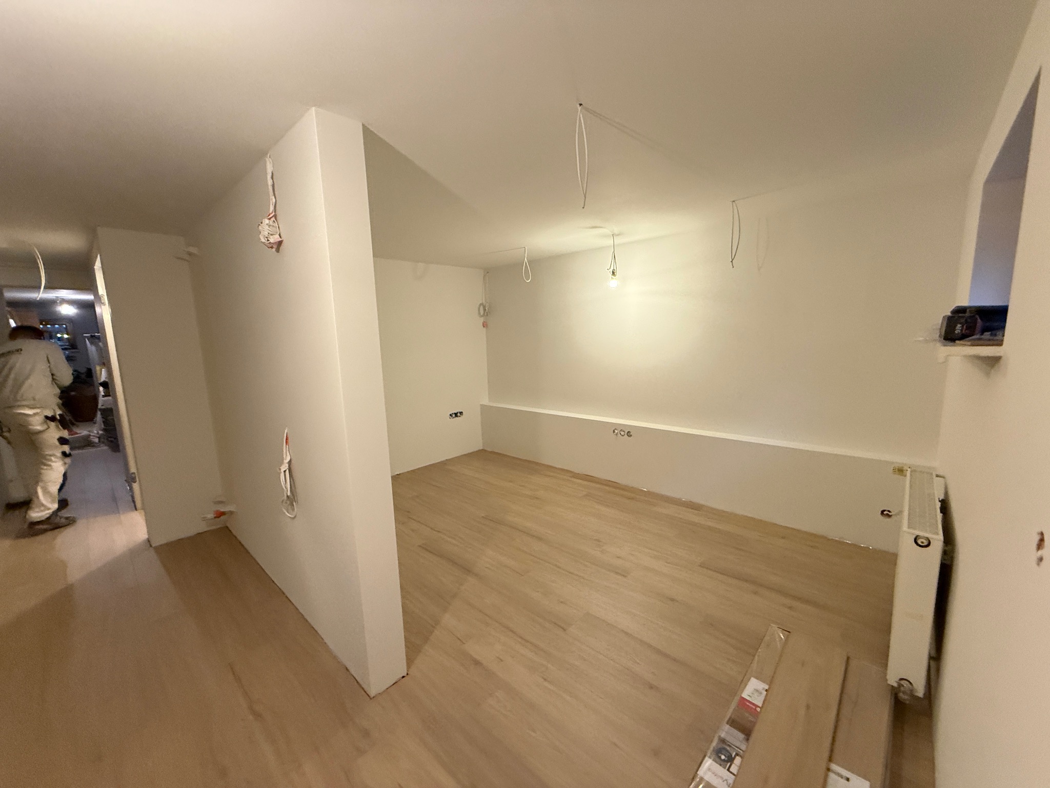 Wnętrze po malowaniu: białe ściany, drewniana podłoga, widoczne kable i instalacje elektryczne. Częściowo widoczny malarz w kombinezonie. Nowoczesne wykończenie.