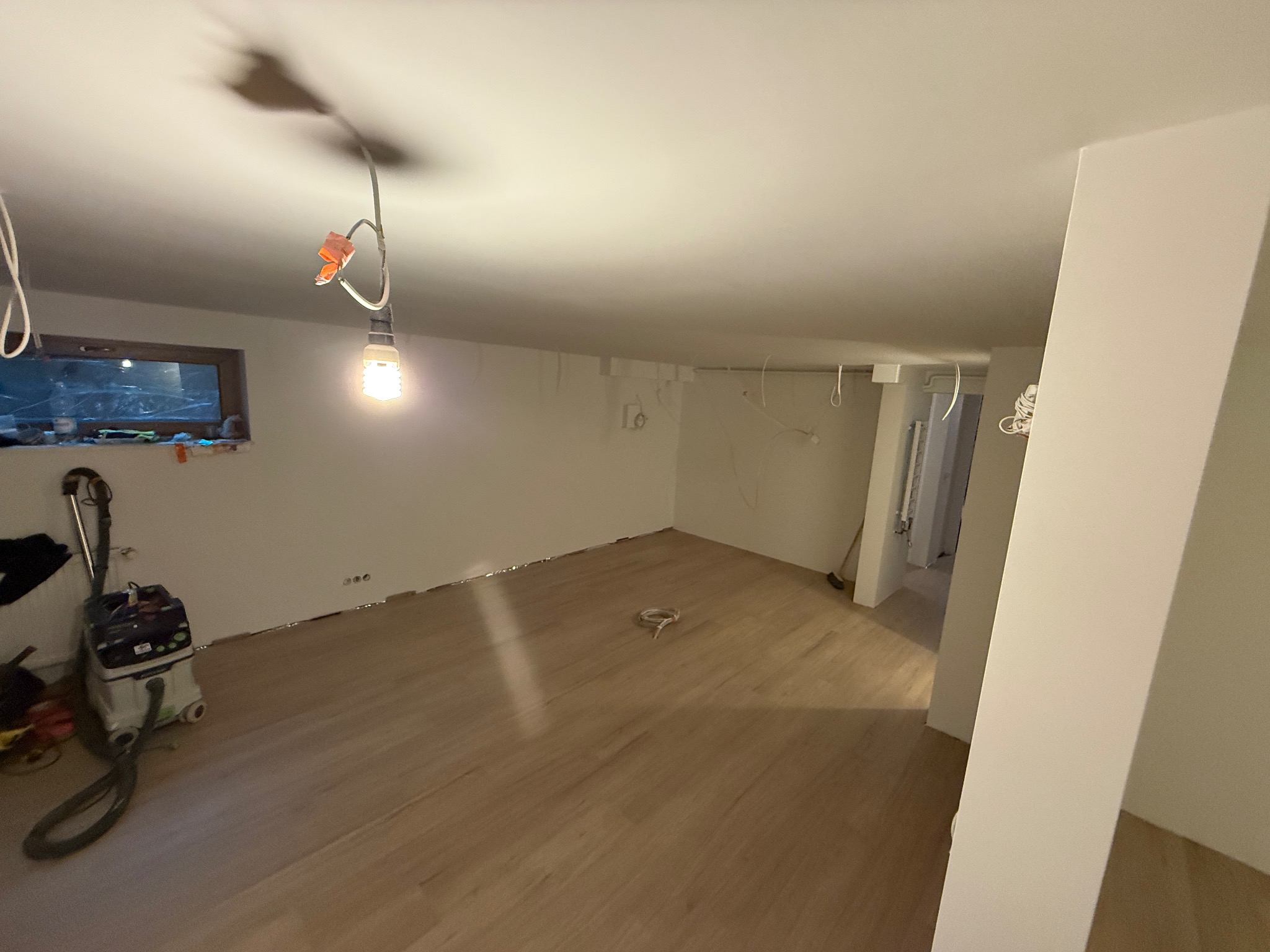 Jasne panele podłogowe w surowym wnętrzu z białymi ścianami. Widoczny odkurzacz przemysłowy, okno i wiszące kable. Etap prac wykończeniowych.