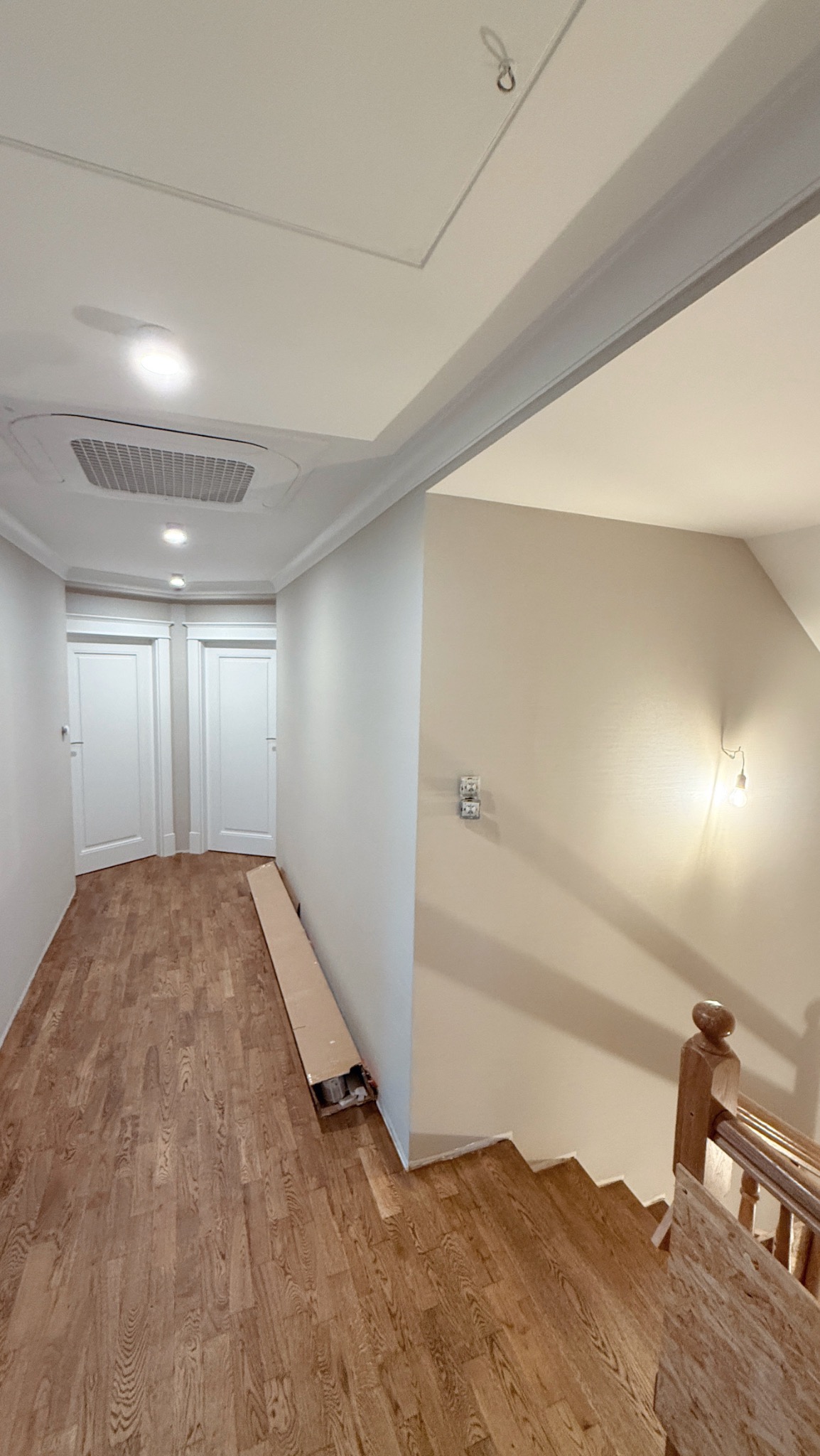Jasne wnętrze po remoncie: świeżo pomalowane ściany w odcieniach beżu i bieli, drewniana podłoga, białe drzwi. Widoczne schody i kartonowe opakowanie przy ścianie.