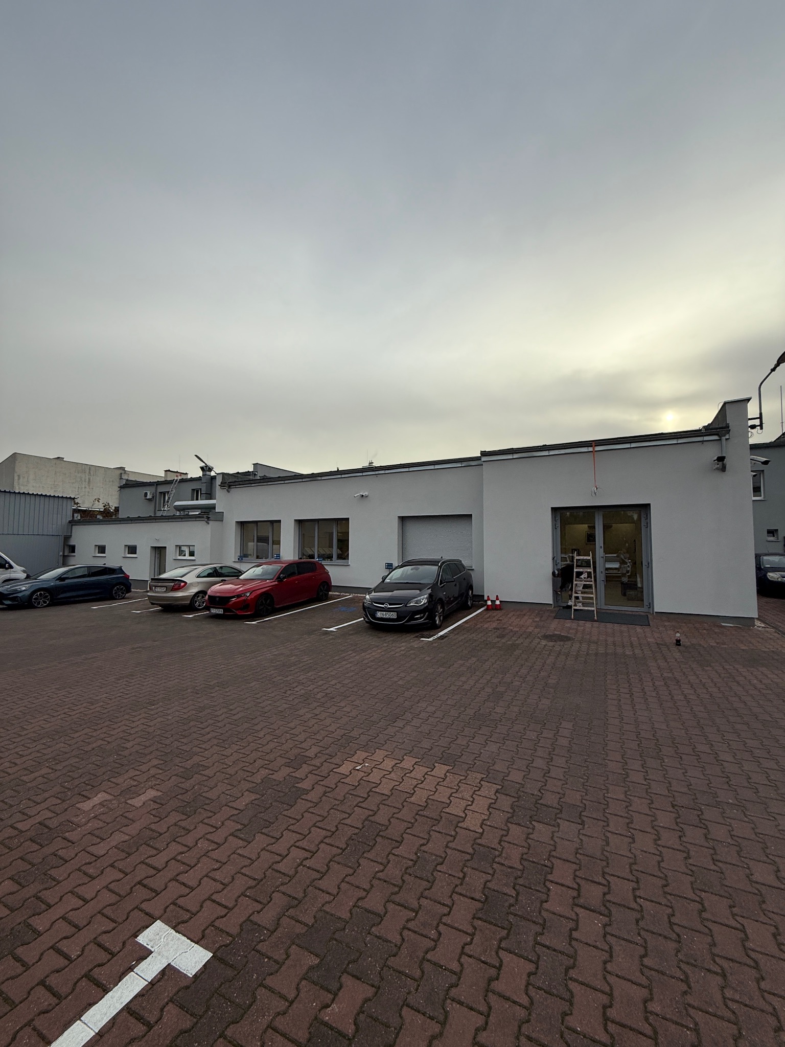 Szary budynek biurowy po malowaniu elewacji, z zaparkowanymi samochodami i drabiną przy wejściu. Widok z zewnątrz, pochmurne niebo.