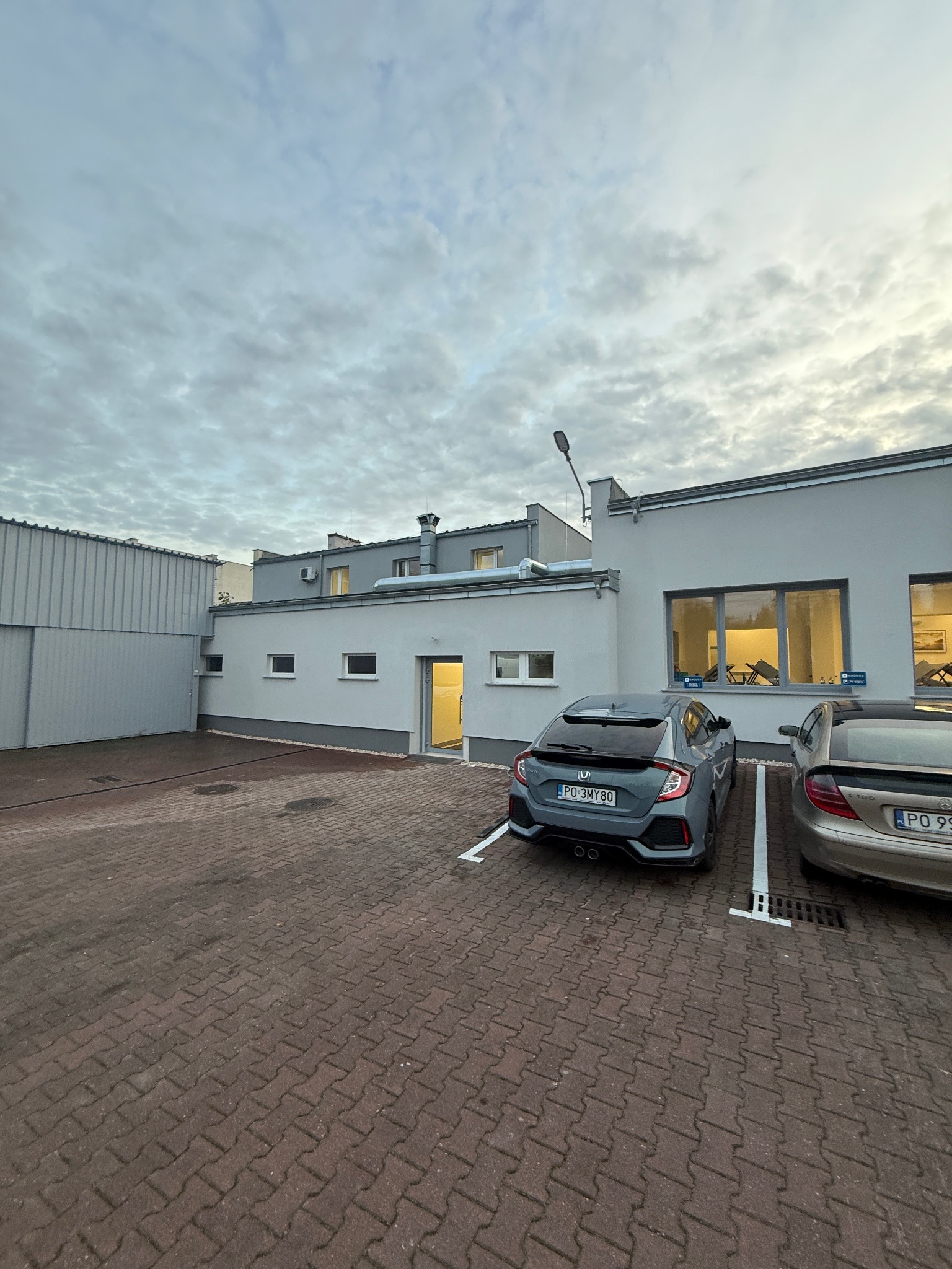 Szary budynek biurowy z zaparkowanymi samochodami na kostce brukowej. Widoczne okna z oświetleniem wewnętrznym i fragment metalowej konstrukcji po lewej stronie.