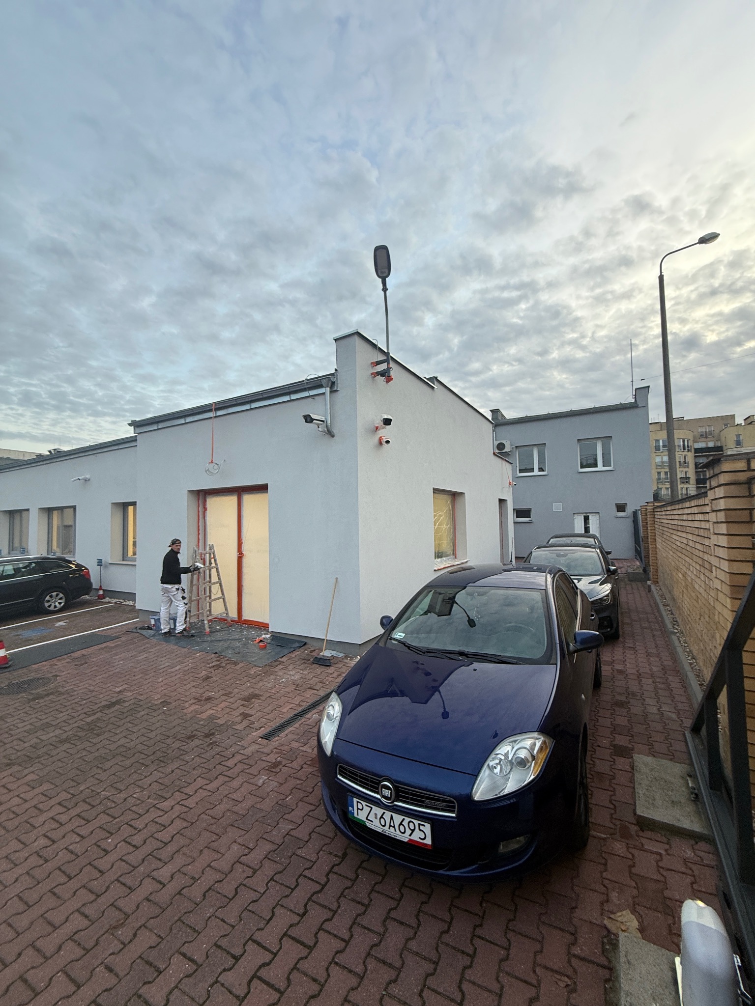 Pracownik na drabinie maluje elewację budynku w kolorze białym. Widoczne zabezpieczone okna i drzwi. Na pierwszym planie zaparkowany samochód.