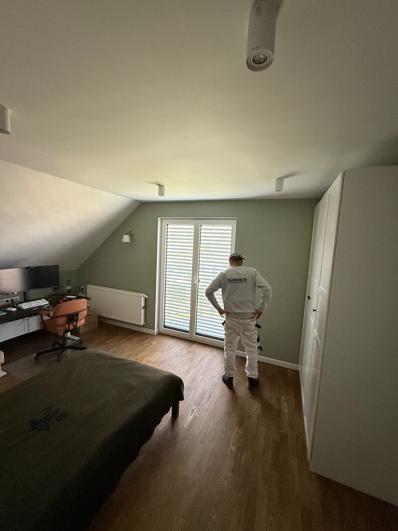 Malowanie ścian w Śremie: Malarz w kombinezonie firmowym stoi w pokoju z zielonymi ścianami, po remoncie. Widoczne meble i okno z żaluzjami.