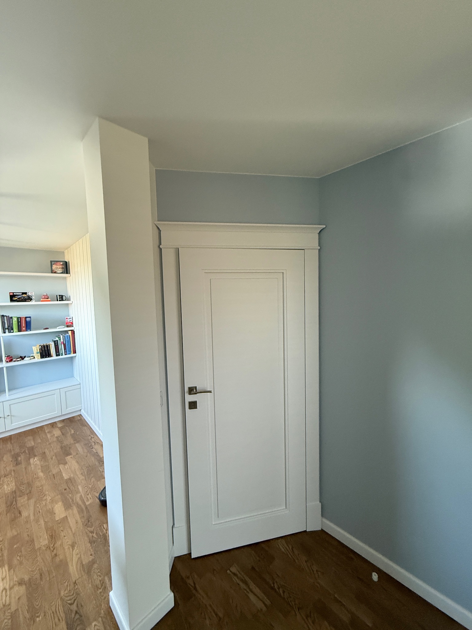 Białe drzwi wewnętrzne z ozdobną ramą w odcieniach błękitu, widoczne z perspektywy korytarza z drewnianą podłogą i regałem na książki w tle. Jasne, naturalne światło.