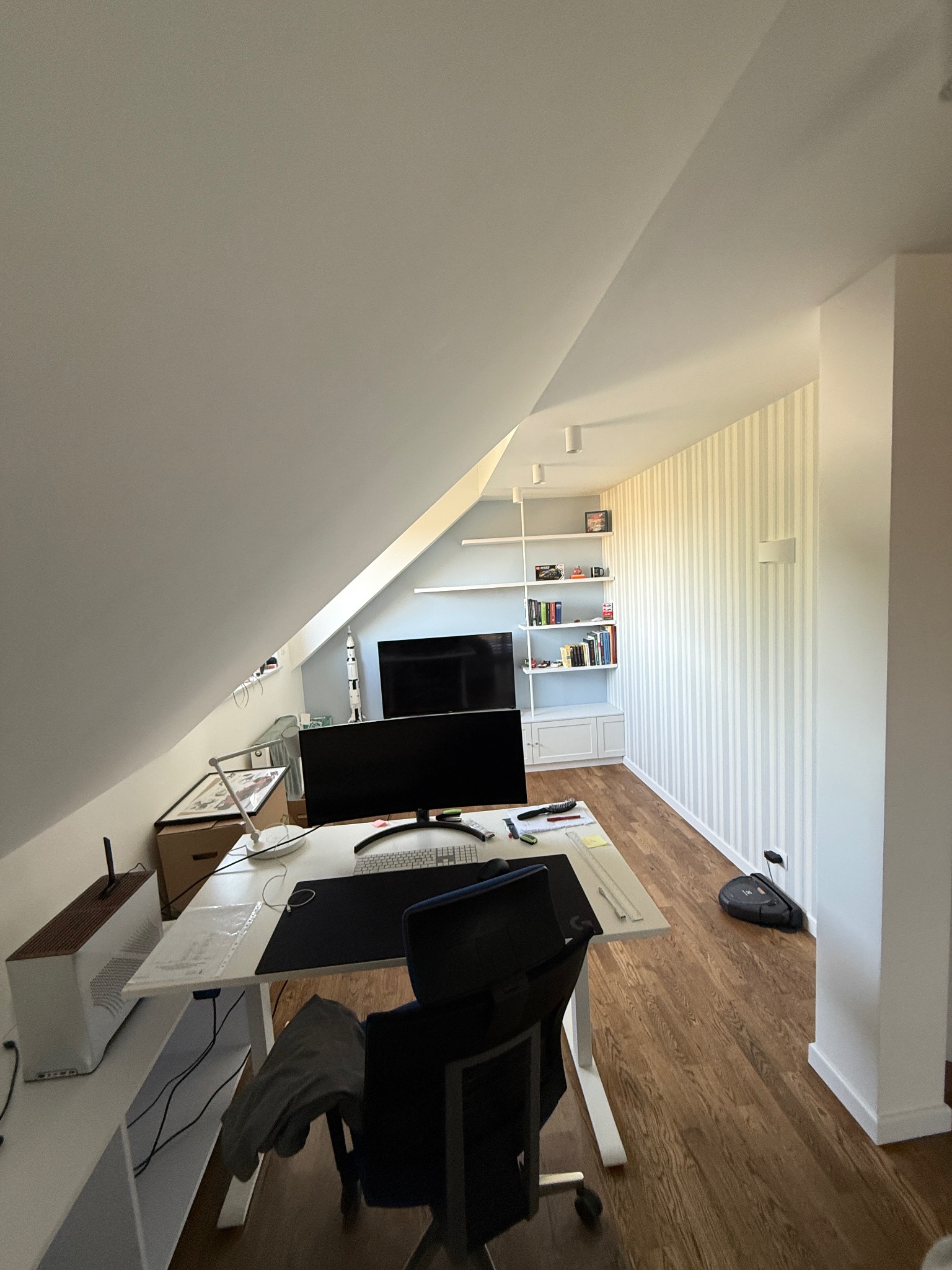 Pokój na poddaszu ze skośnym sufitem, biurkiem z komputerem i regałem z książkami. Ściana w biało-szare pasy. Podłoga drewniana. Widok na uporządkowane wnętrze.