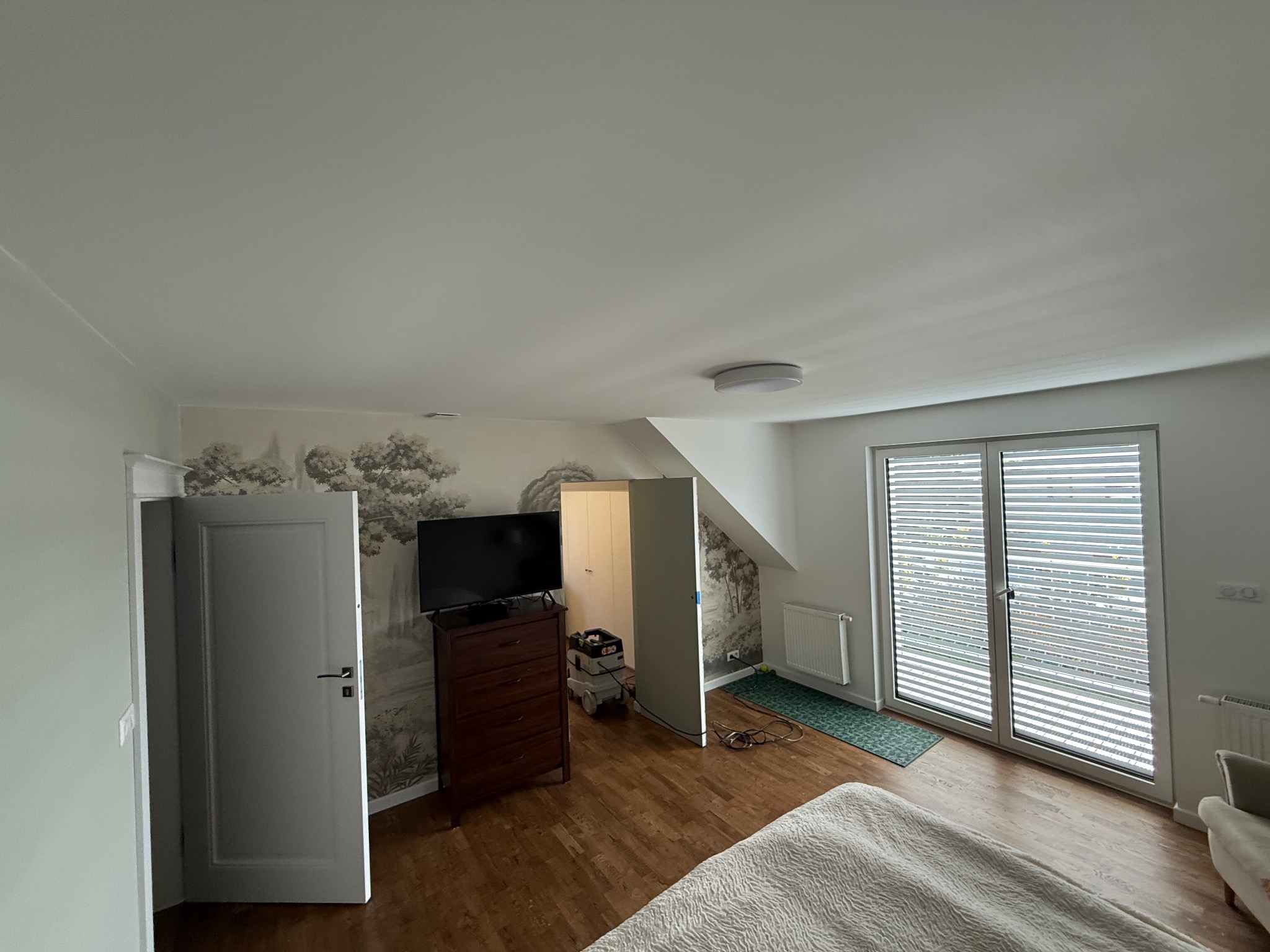 Jasne wnętrze pokoju z drewnianą podłogą, komodą z telewizorem i fototapetą z motywem drzew. Widoczne okno balkonowe z żaluzjami i otwarte drzwi.