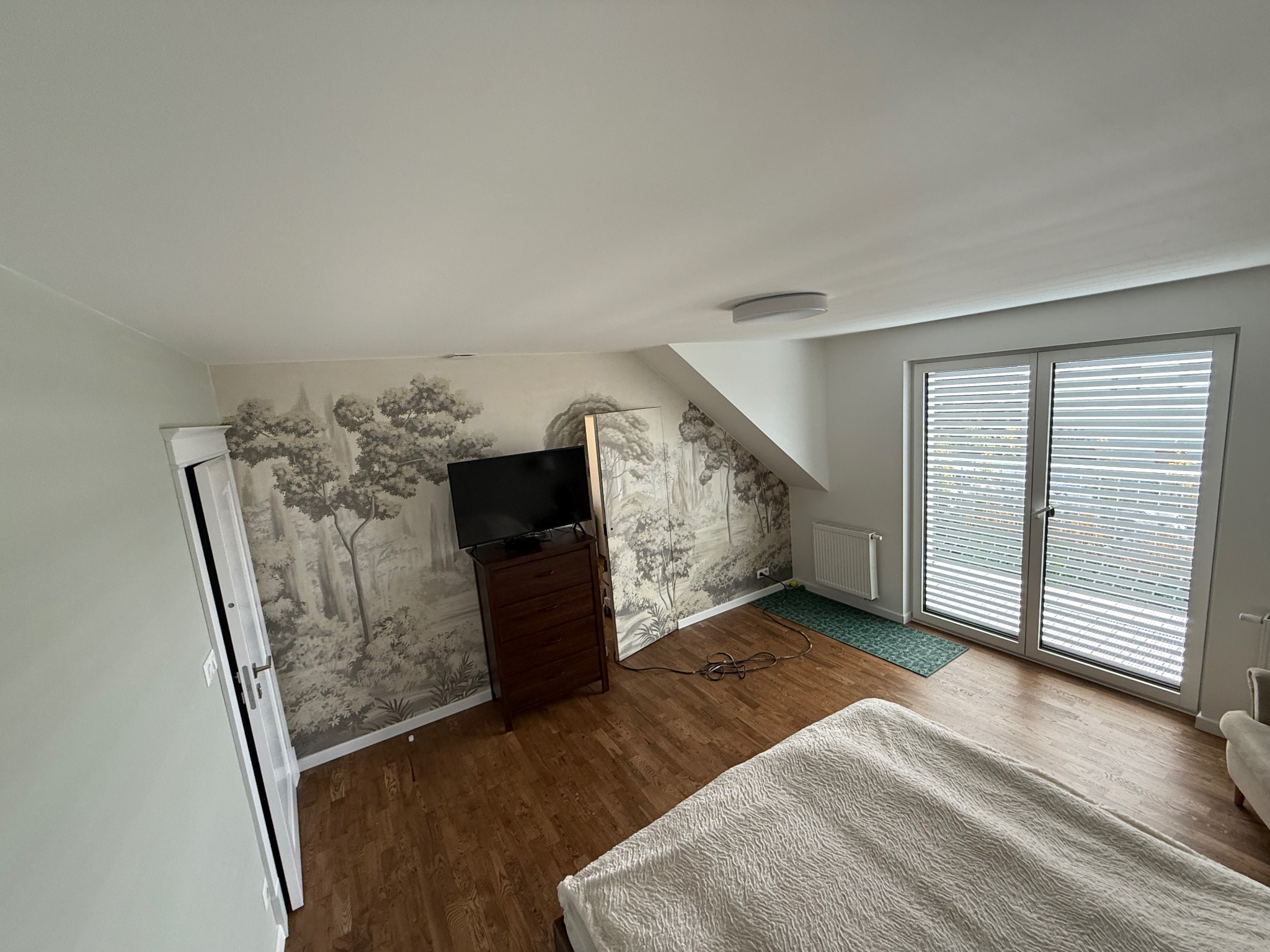 Pokój na poddaszu z tapetą w motywy leśne, drewnianą podłogą i komodą. Widoczne okno z żaluzjami i fragment łóżka. Ściany pomalowane na jasny odcień szarości.