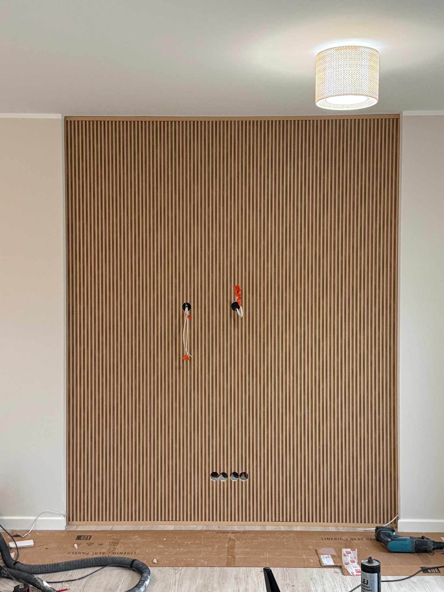 Montaż paneli lamelowych na ścianie z wyprowadzonymi kablami elektrycznymi i gniazdkami. Widoczne narzędzia i akcesoria montażowe. Oświetlenie sufitowe.