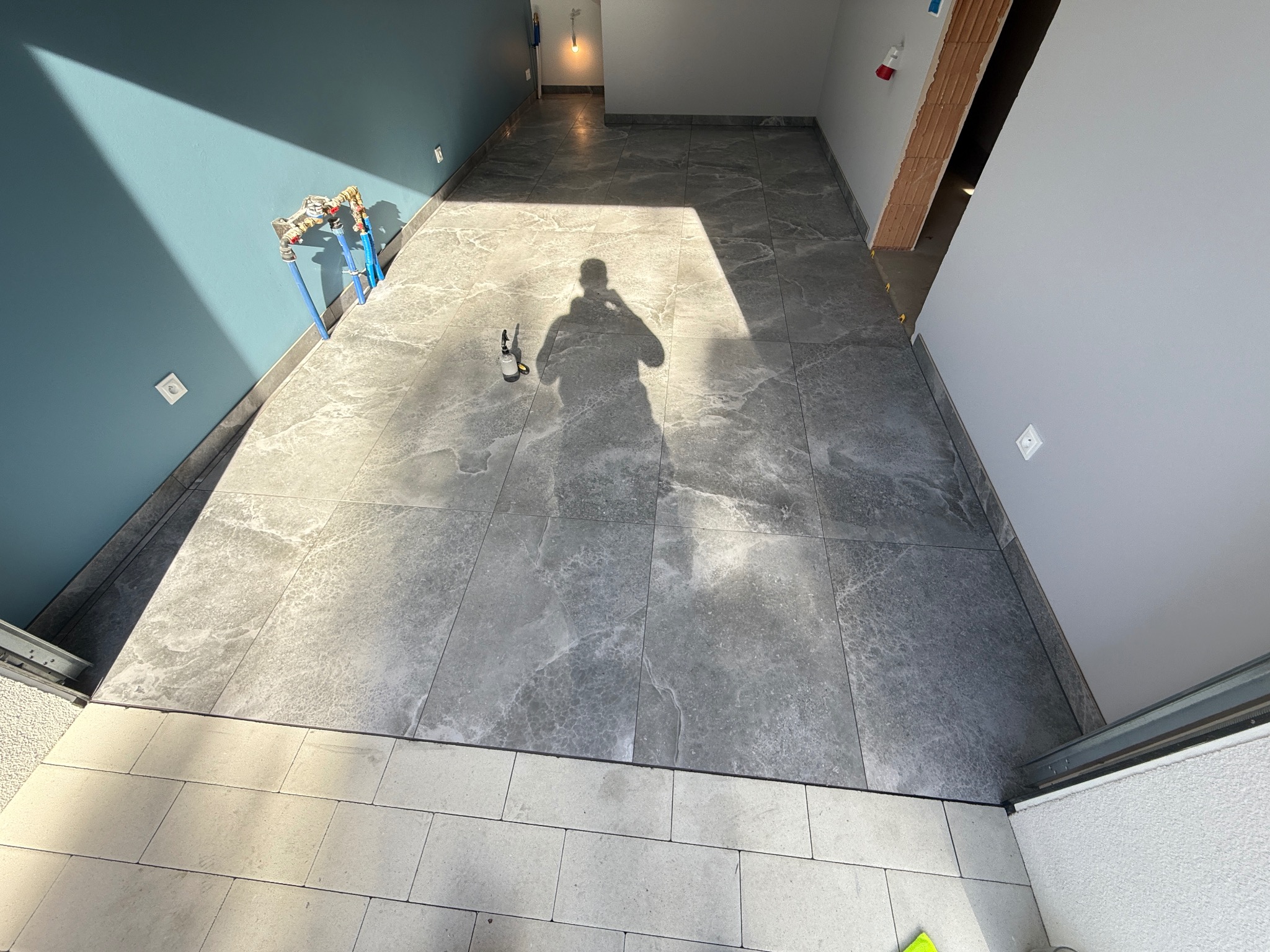 Wykończone wnętrze z szarymi płytkami podłogowymi imitującymi marmur, błękitną ścianą i instalacją hydrauliczną. Widoczny cień osoby robiącej zdjęcie.