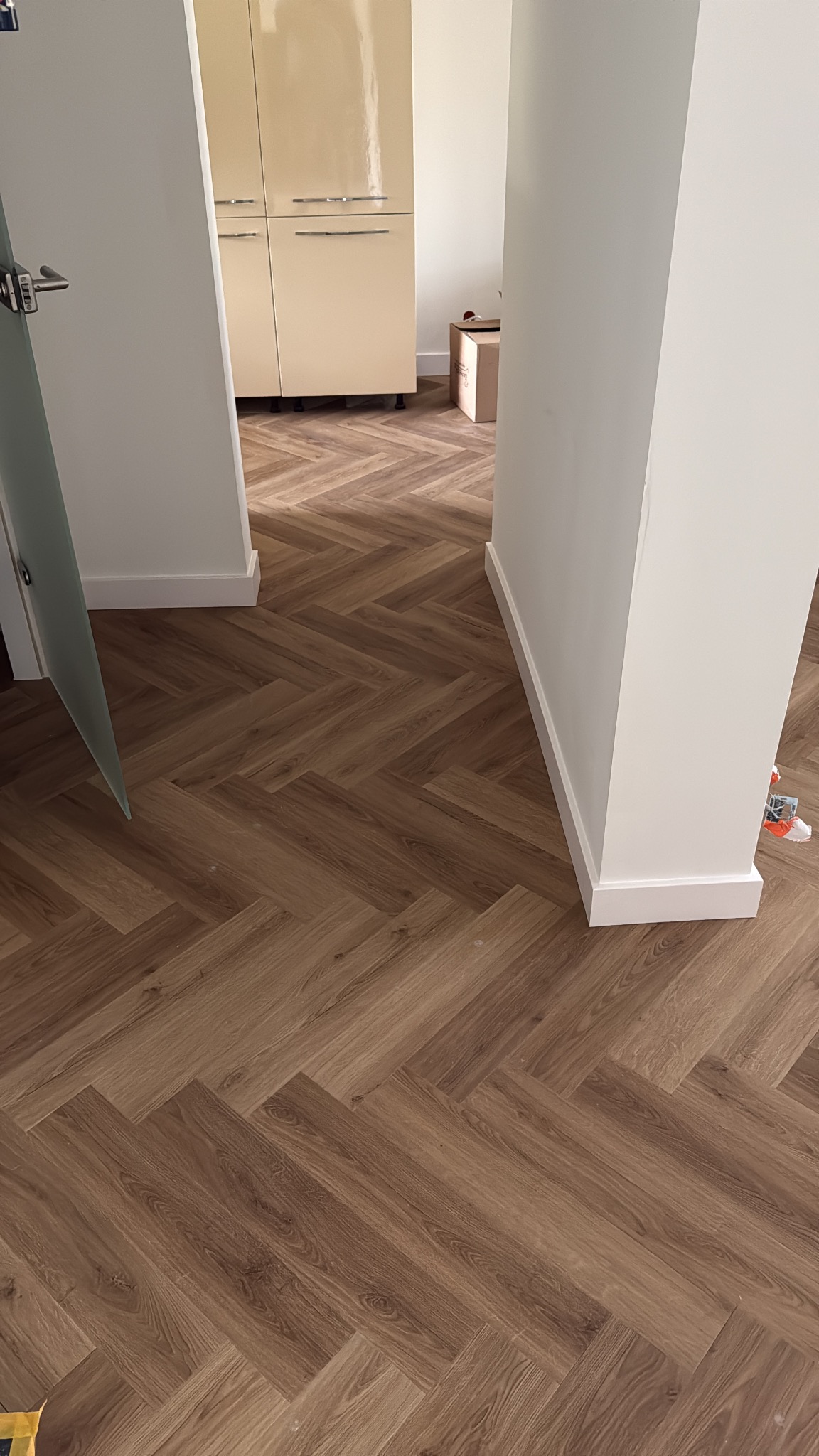 Ukończona podłoga z paneli ułożonych w jodełkę, widok z perspektywy podłogi, białe listwy przypodłogowe, fragment drzwi i szafki w tle. Jasne wnętrze.