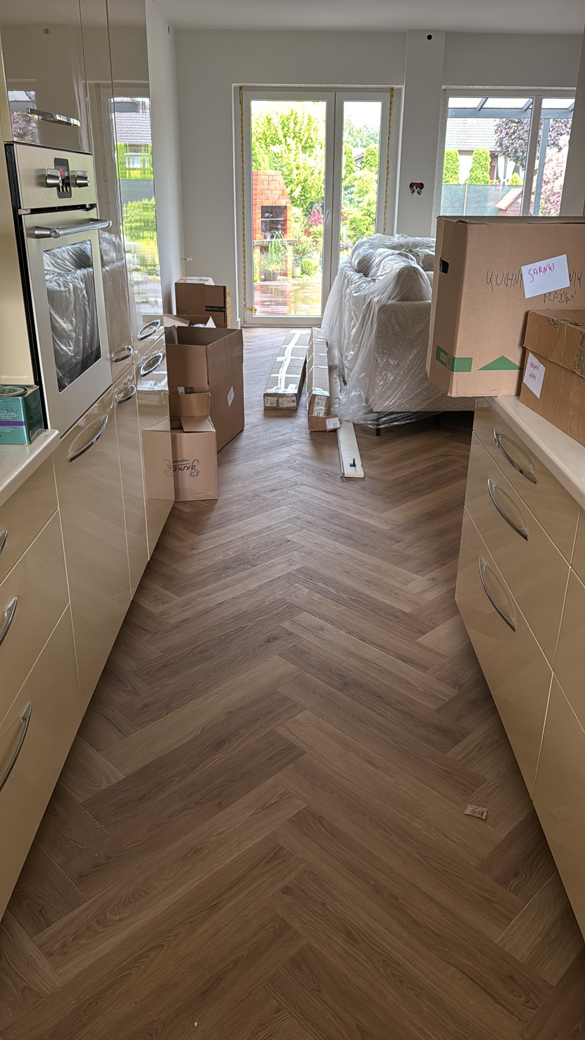 Ukończona podłoga w jodełkę w jasnej kuchni z beżowymi szafkami. Widoczne kartony i zabezpieczone meble, sugerujące świeżo zakończony remont.