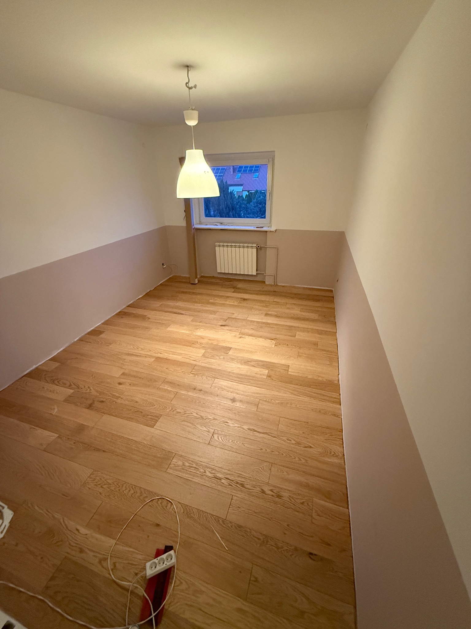 Pokój po malowaniu ścian na dwa kolory: górna część biała, dolna w odcieniach beżu. Na podłodze parkiet. Widoczne gniazdko elektryczne i lampa sufitowa.