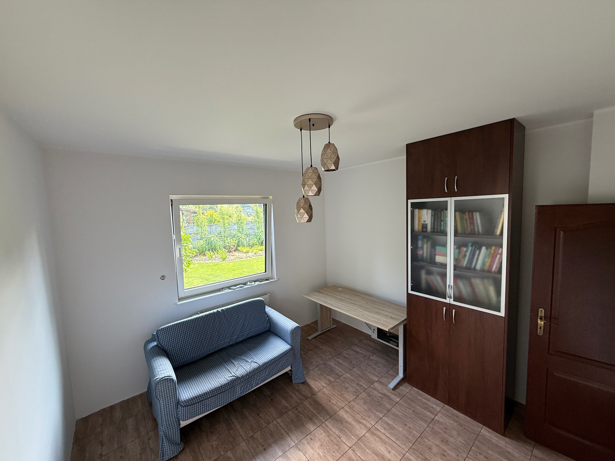 Jasne wnętrze pokoju z sofą w kratę, biurkiem i regałem na książki. Widoczne okno z widokiem na zieleń. Lampa wisząca z trzema kloszami. Podłoga wyłożona płytkami imitującymi drewno.