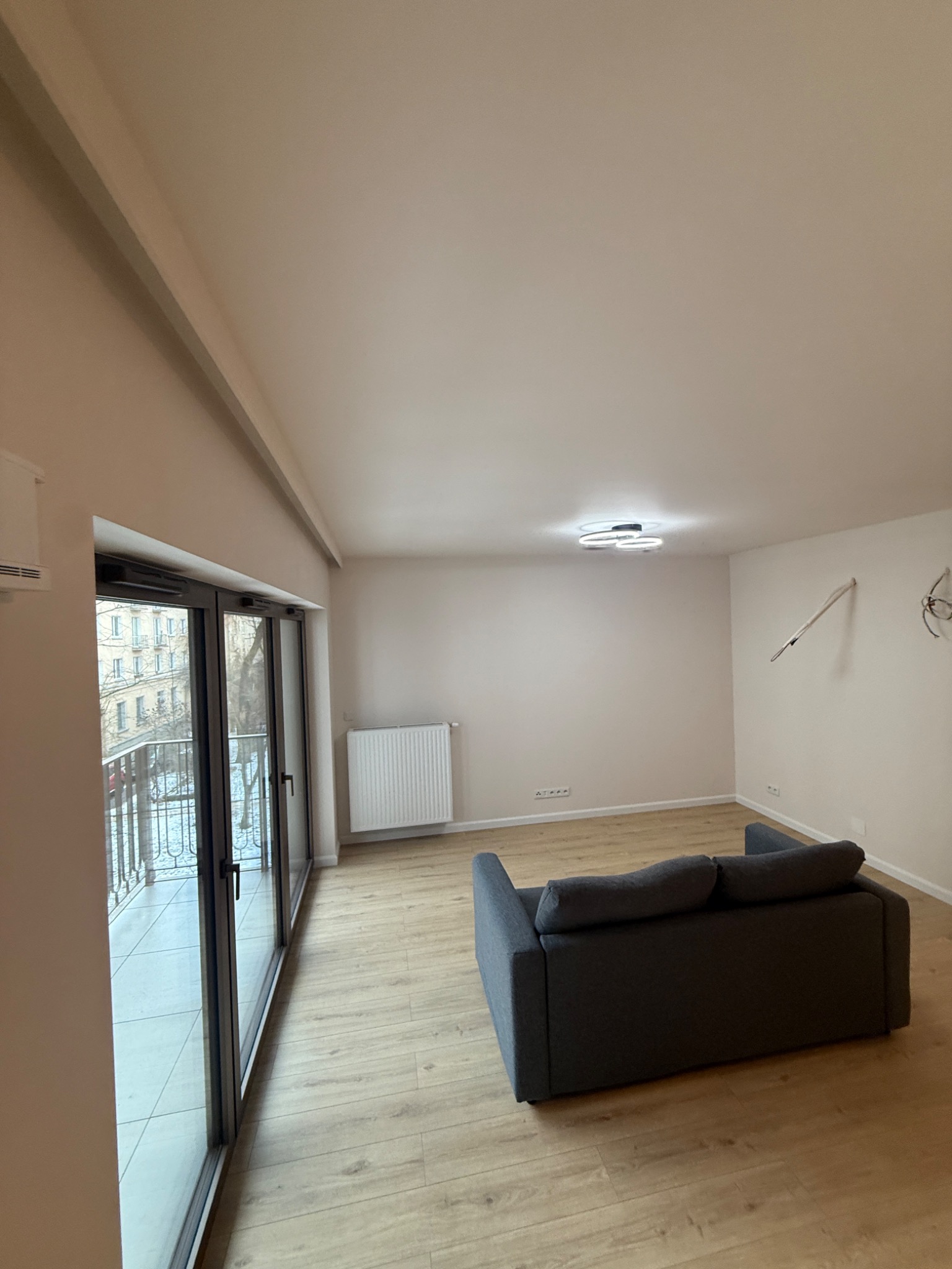 Jasne wnętrze po remoncie: parkiet, szara sofa, okno balkonowe z widokiem na miasto. Na ścianie widoczne niedokończone instalacje elektryczne.