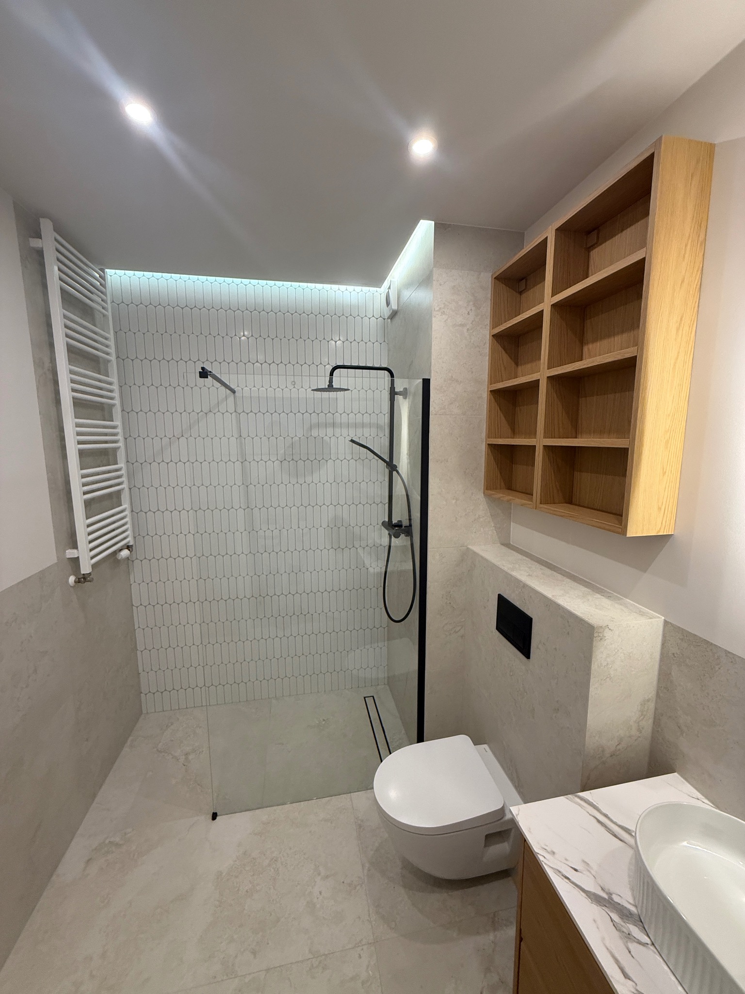 Nowoczesna łazienka po remoncie: kabina walk-in z czarną armaturą, białe płytki w kształcie łezki, drewniana szafka i umywalka nablatowa. Minimalistyczny design.
