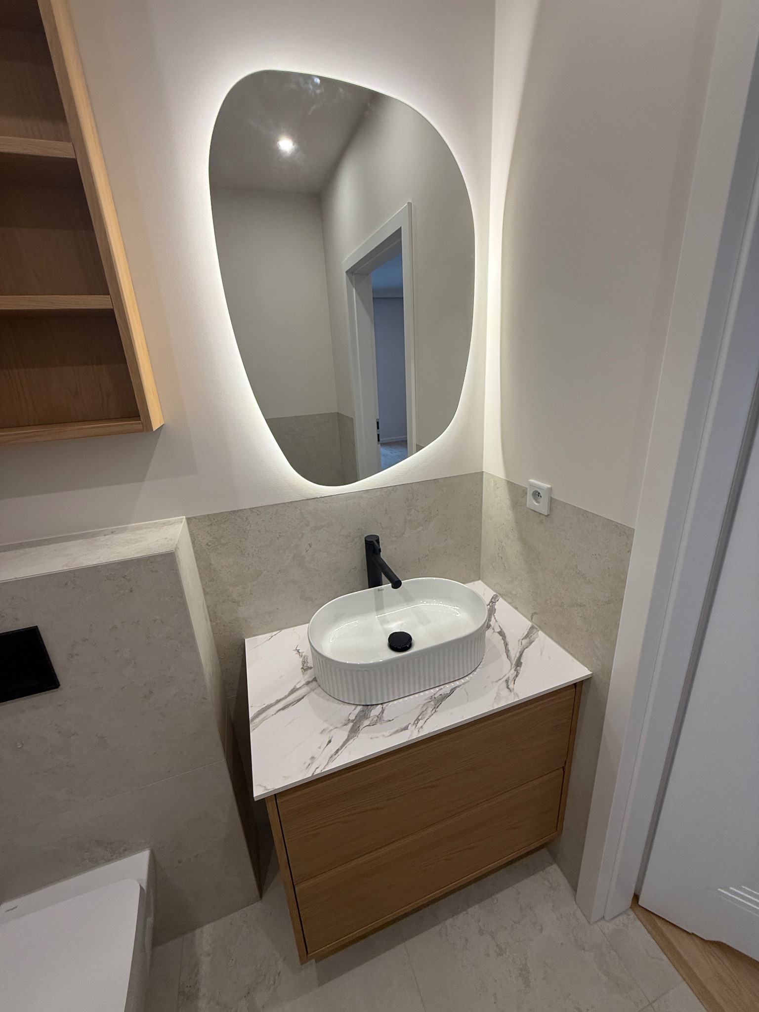 Nowoczesna łazienka po remoncie: owalne lustro z podświetleniem LED, biała umywalka nablatowa na drewnianej szafce z marmurowym blatem, szare płytki.