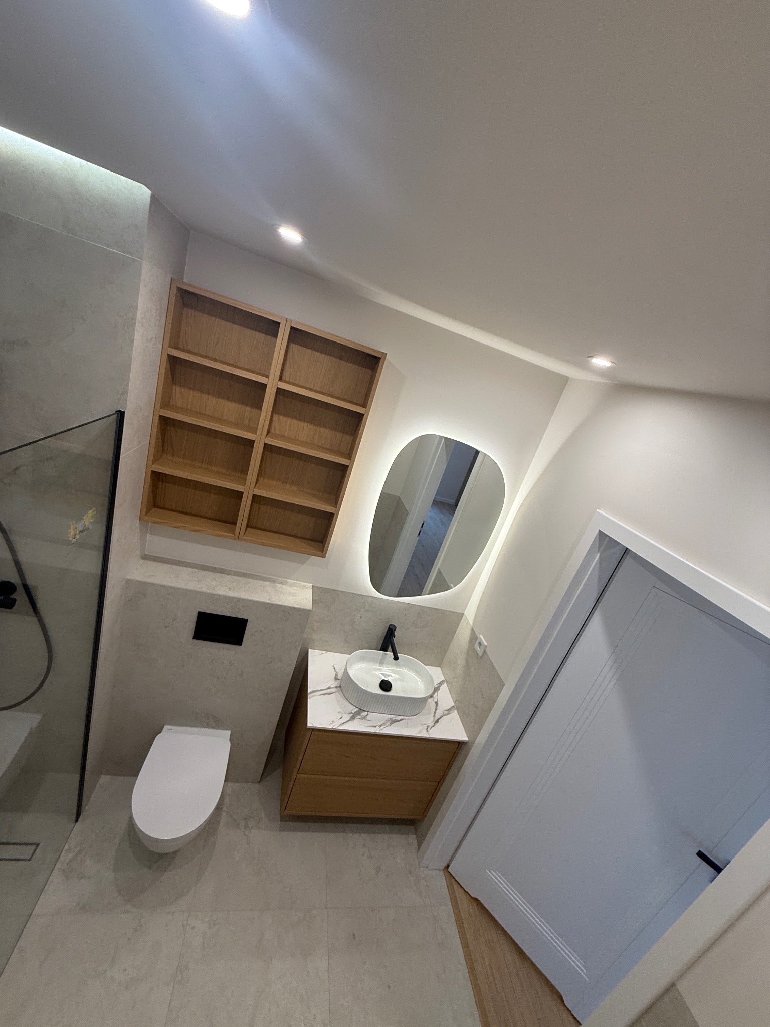 Nowoczesna łazienka po remoncie: biała ceramika, drewniane szafki, podświetlane lustro o nieregularnym kształcie i szklana kabina prysznicowa. Minimalistyczny design.