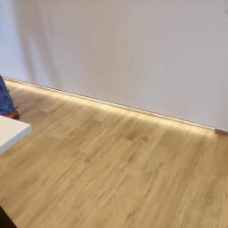 SAJ-BUD - Nowoczesne wnętrze z podświetlaną listwą przypodłogową LED, jasnymi panelami podłogowymi i fragmentem białej ściany. Widoczny fragment osoby w ubraniu roboczym.