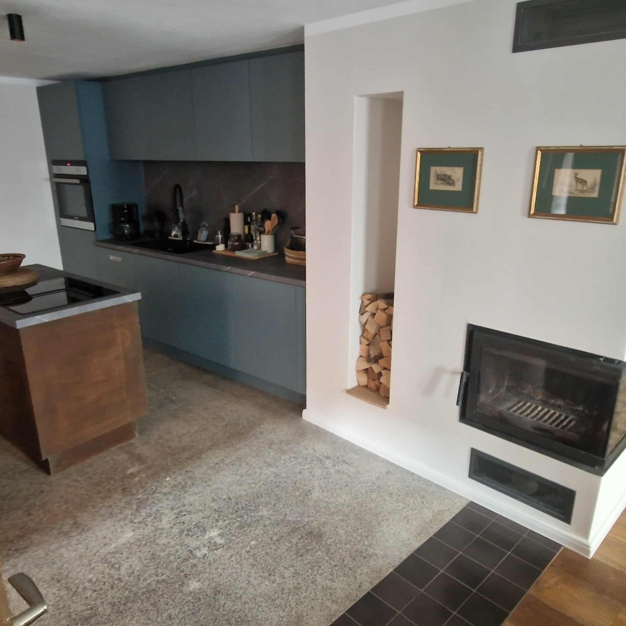 Nowoczesna kuchnia z szarymi szafkami, wyspą z drewnianym blatem i kominkiem z wnęką na drewno. Dwa obrazy w złotych ramach wiszą nad kominkiem.