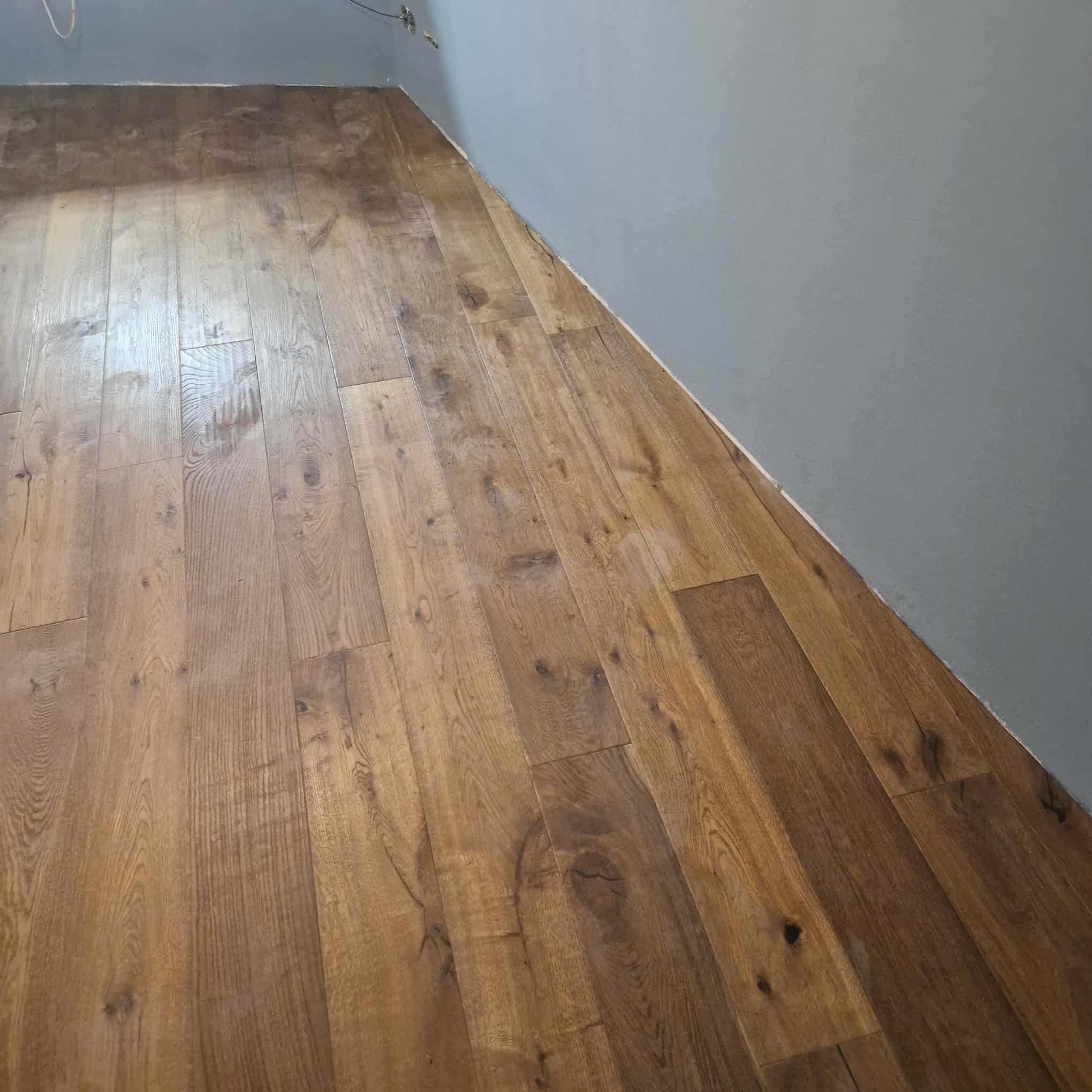 Ułożona podłoga z paneli drewnopodobnych w odcieniach brązu, widoczna przy szarej ścianie. Układ paneli wzdłuż ściany, widoczne detale struktury drewna.