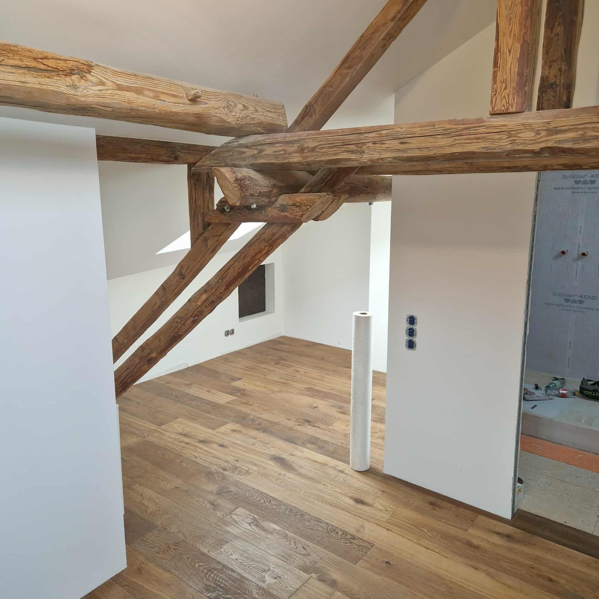 Jasne wnętrze po remoncie: drewniane belki stropowe, parkiet w jodełkę, świeżo pomalowane ściany. Widoczny fragment łazienki w trakcie wykończenia.